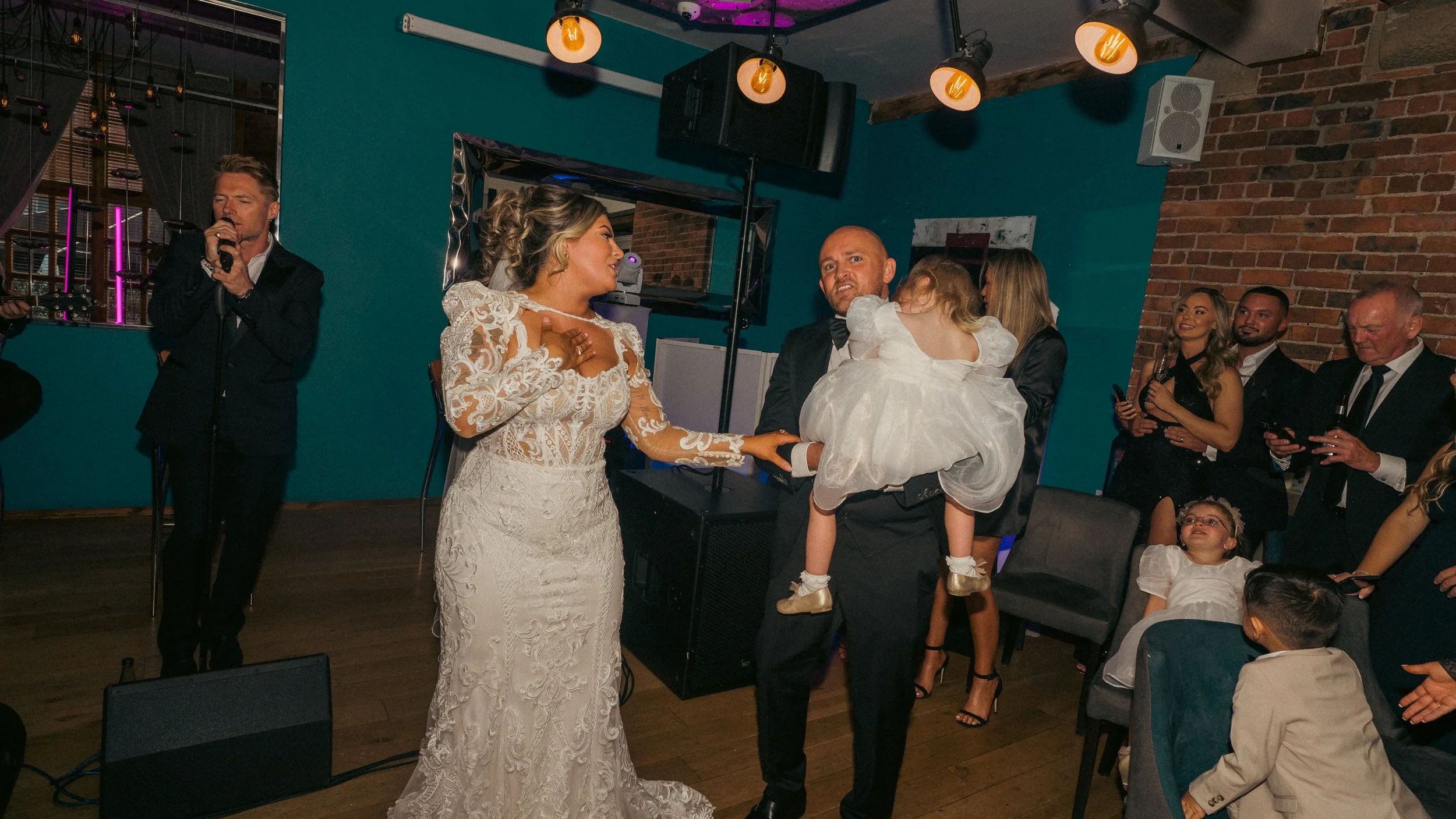 A wedding celebration with the bride in a white lace gown and the groom holding a little girl in a white dress, surrounded by wedding guests in formal attire, some looking at the couple, in a decorated indoor venue with exposed brick walls and warm l