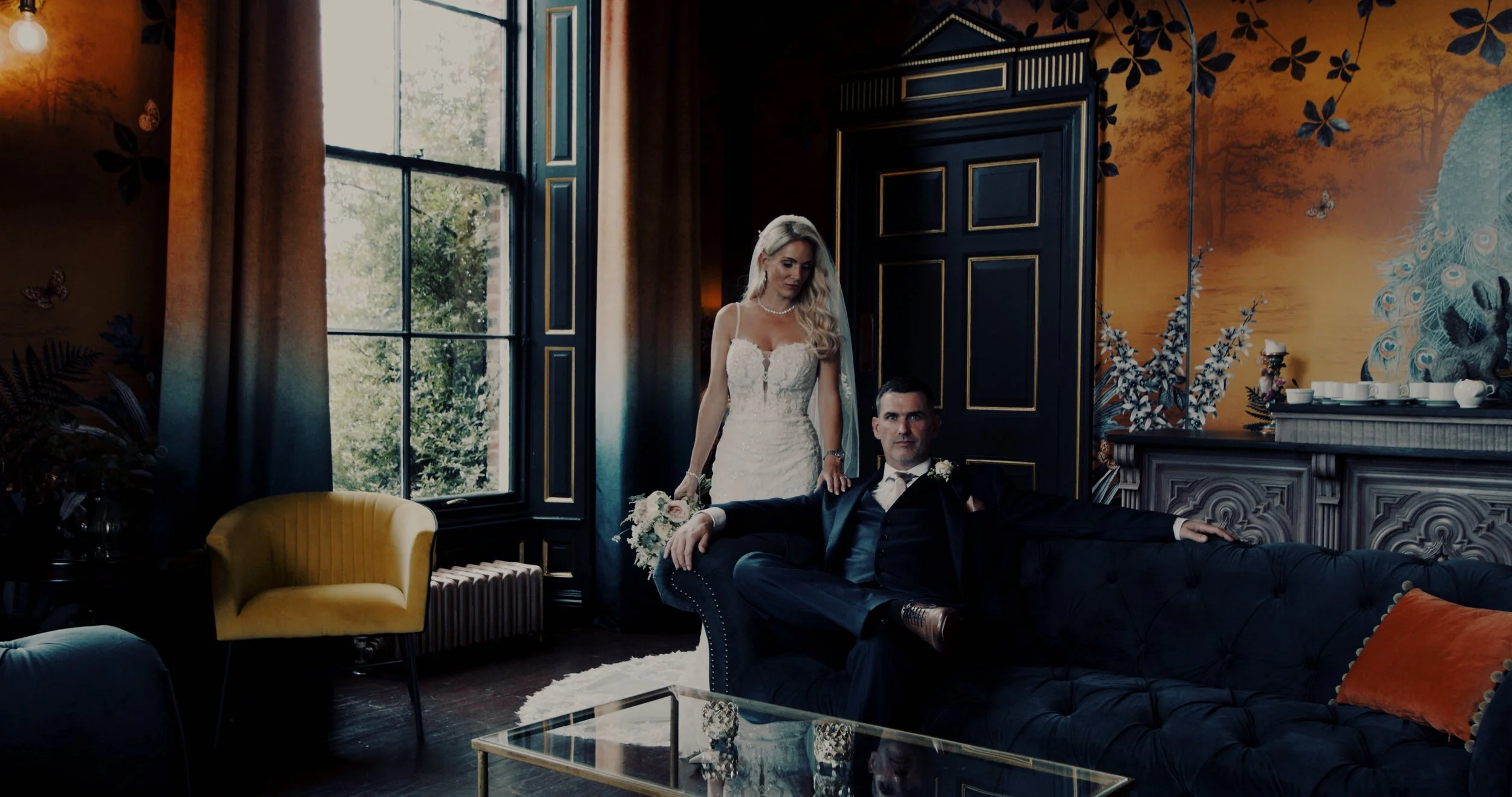 A bride and groom in a decorated room with a black tufted sofa, child yellow chair, and a painted wall with nature and peacock motifs. The bride in a white wedding dress stands behind the groom who is seated on the sofa.