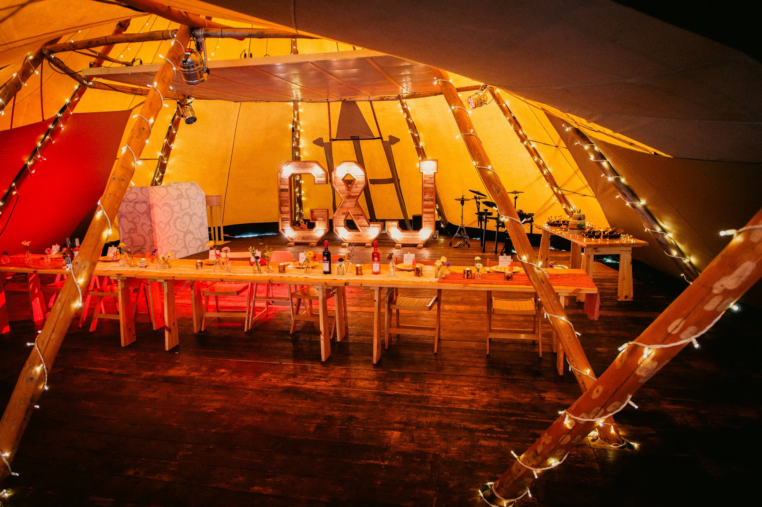 Decorated interior of a wooden tent with a long dining table set with bottles, small flower arrangements, and placards, illuminated with string lights. Large illuminated letters spelling 'G&L' and musical instruments are visible in the background.