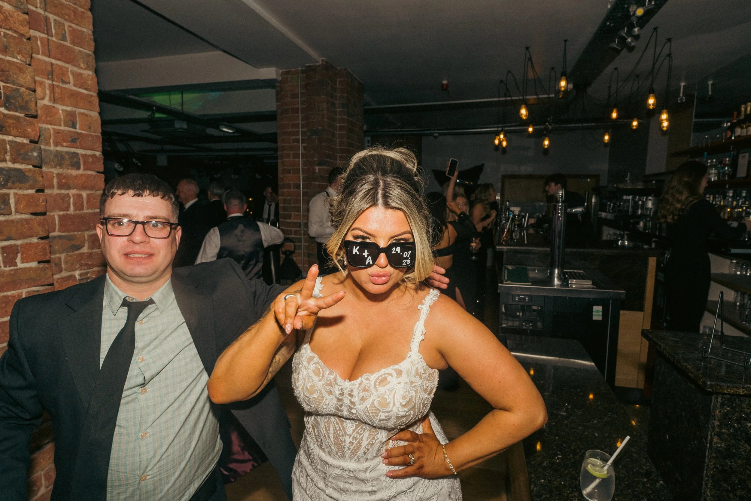 A woman in a white lace dress and sunglasses with custom writing poses with a man in a black suit and glasses at a lively bar or club. The woman is making a peace sign with her fingers. There are other people in the background enjoying the night.