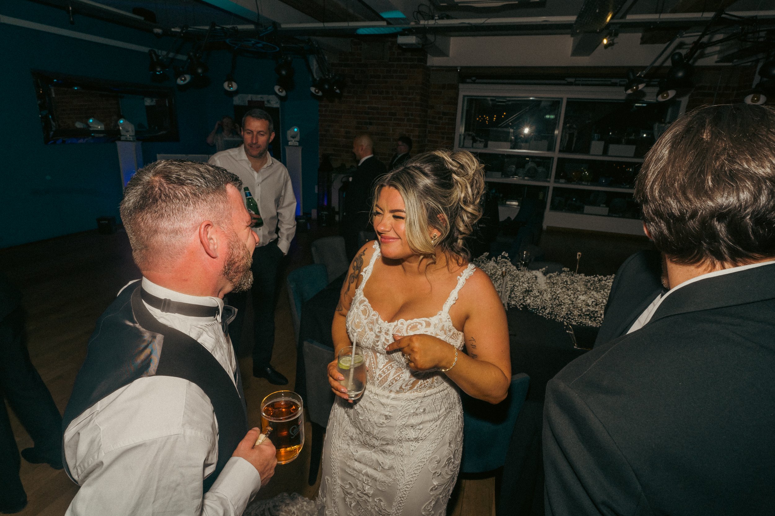 A bride in a white lace wedding dress holding a drink, smiling and talking to a man in formal attire with a drink in his hand, at a wedding reception with other guests in the background.
