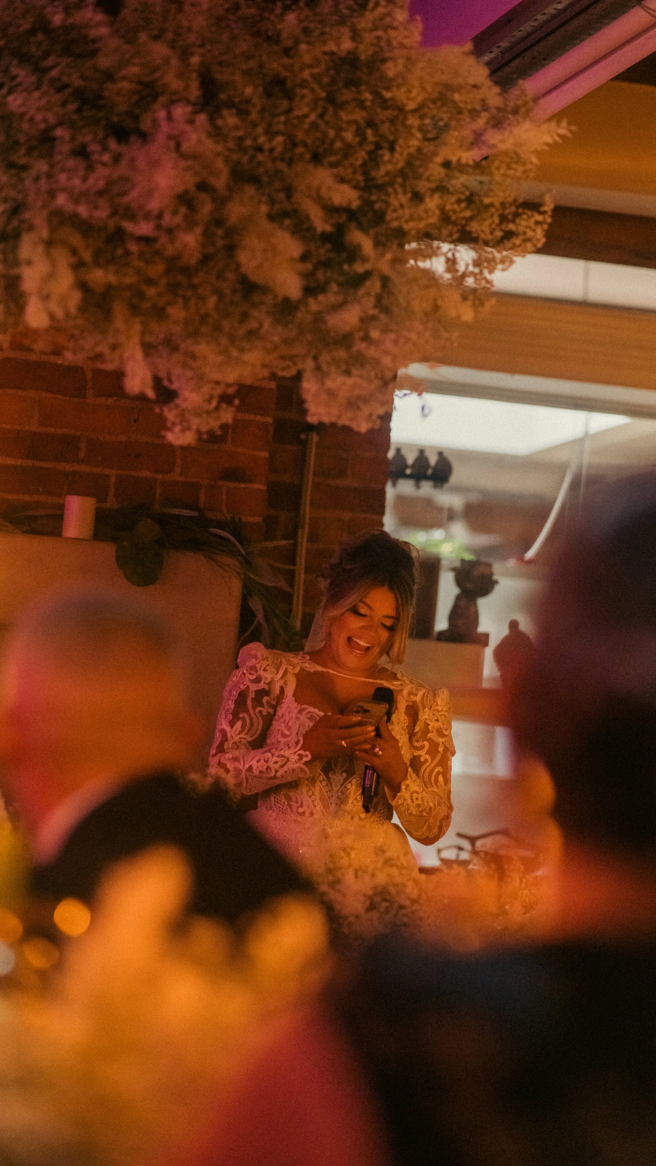 A woman in a lace dress is smiling and looking at her phone, standing indoors near a brick wall with dim, pinkish lighting, surrounded by flowers and decorative objects.