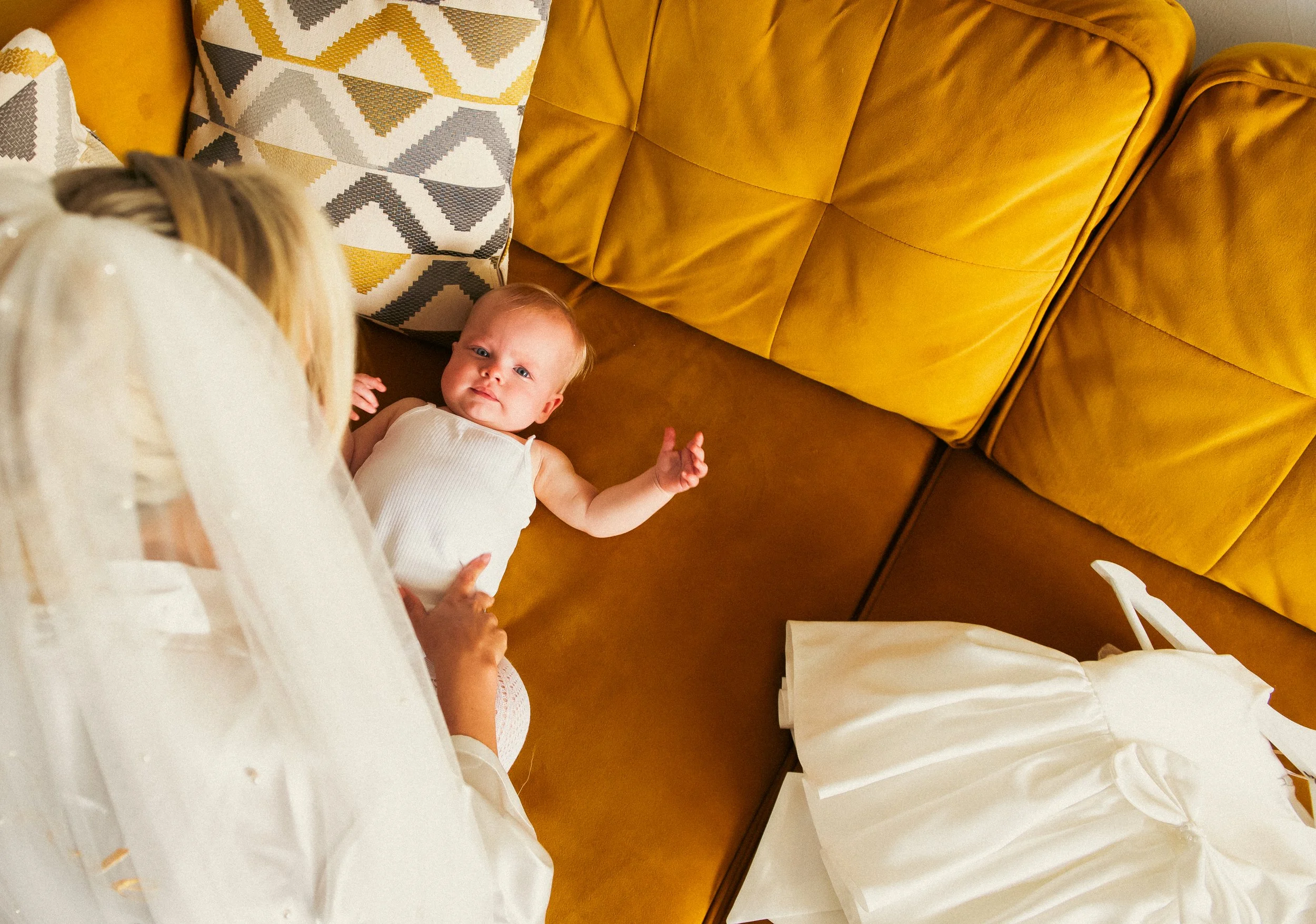 A baby lying on a brown couch with mustard and patterned pillows nearby, viewed from above, while an adult with blond hair in a white outfit interacts with the baby.