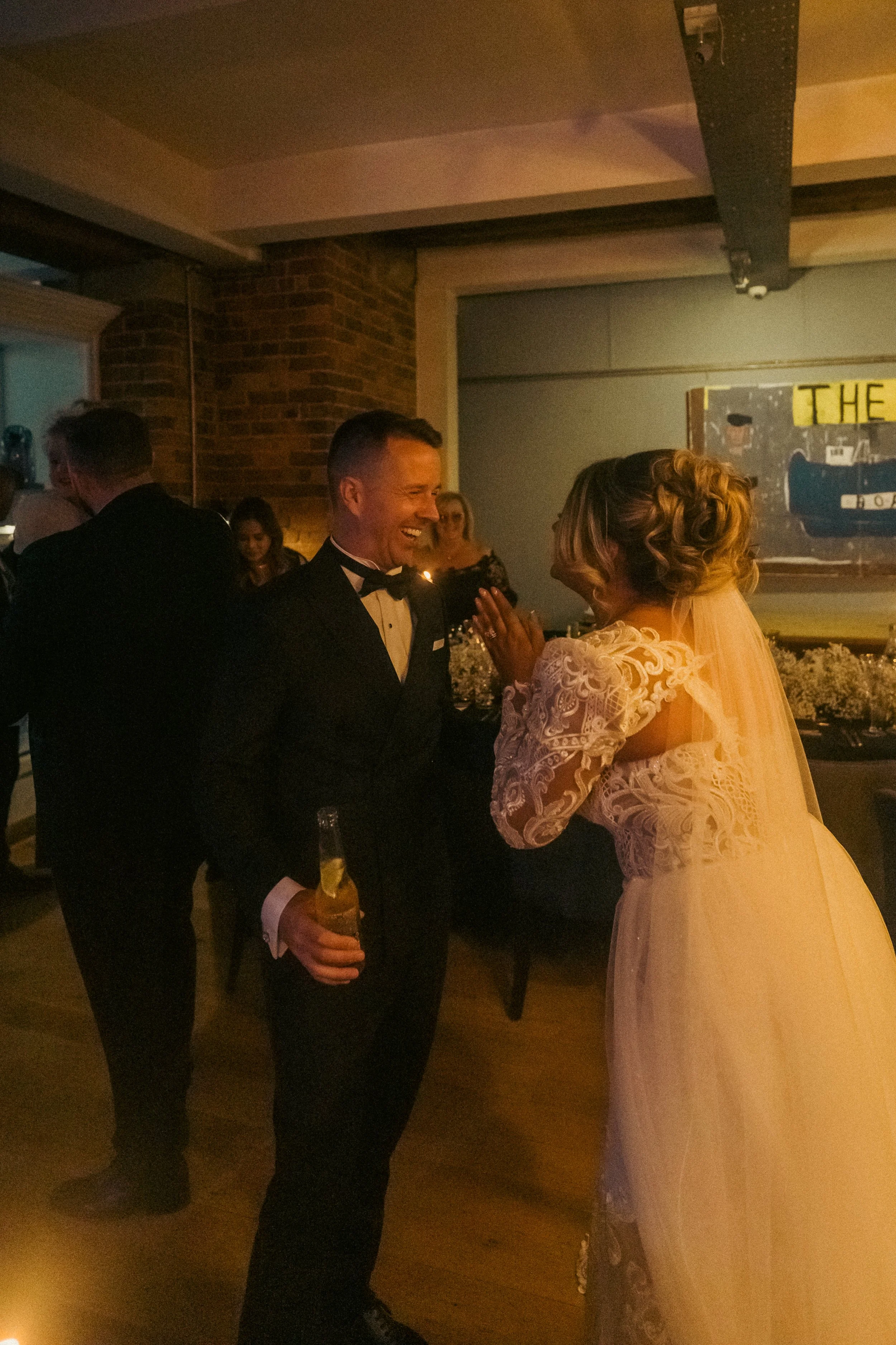 A man and a woman in wedding attire share a joyful conversation at a wedding reception, with other guests in the background and dim lighting.