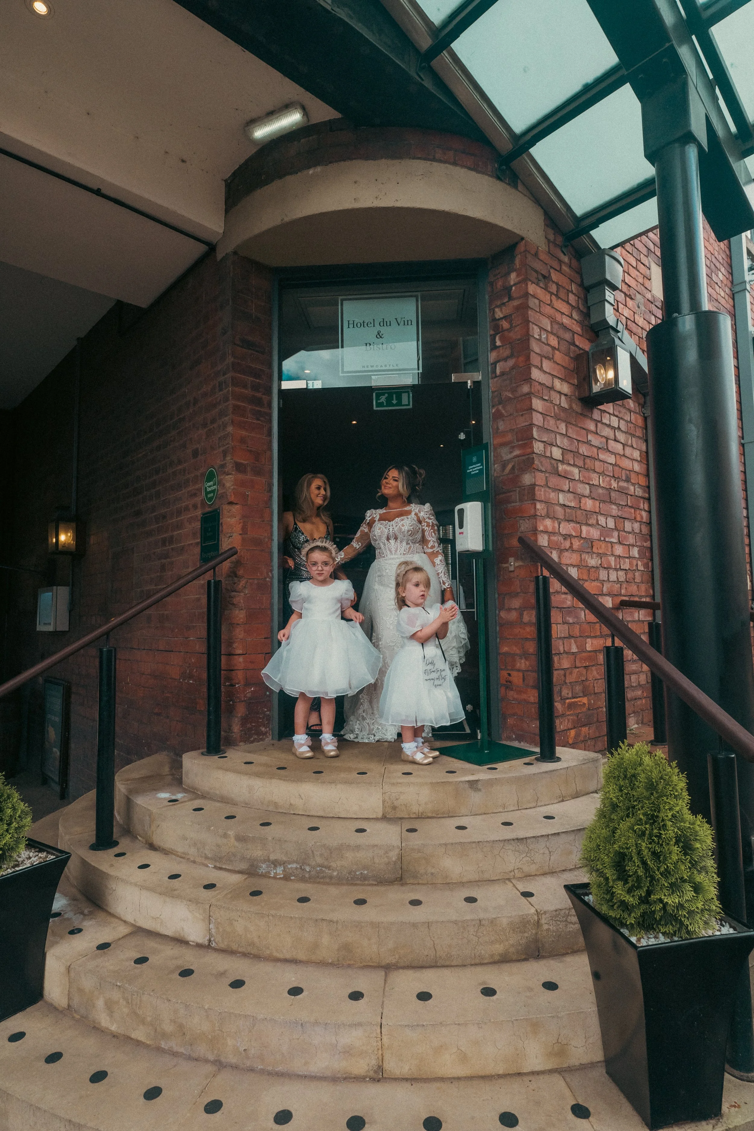 Four women standing on a doorway step. Two young girls in white dresses and two women, one in a wedding gown and the other in a black dress, at the entrance of a brick building.