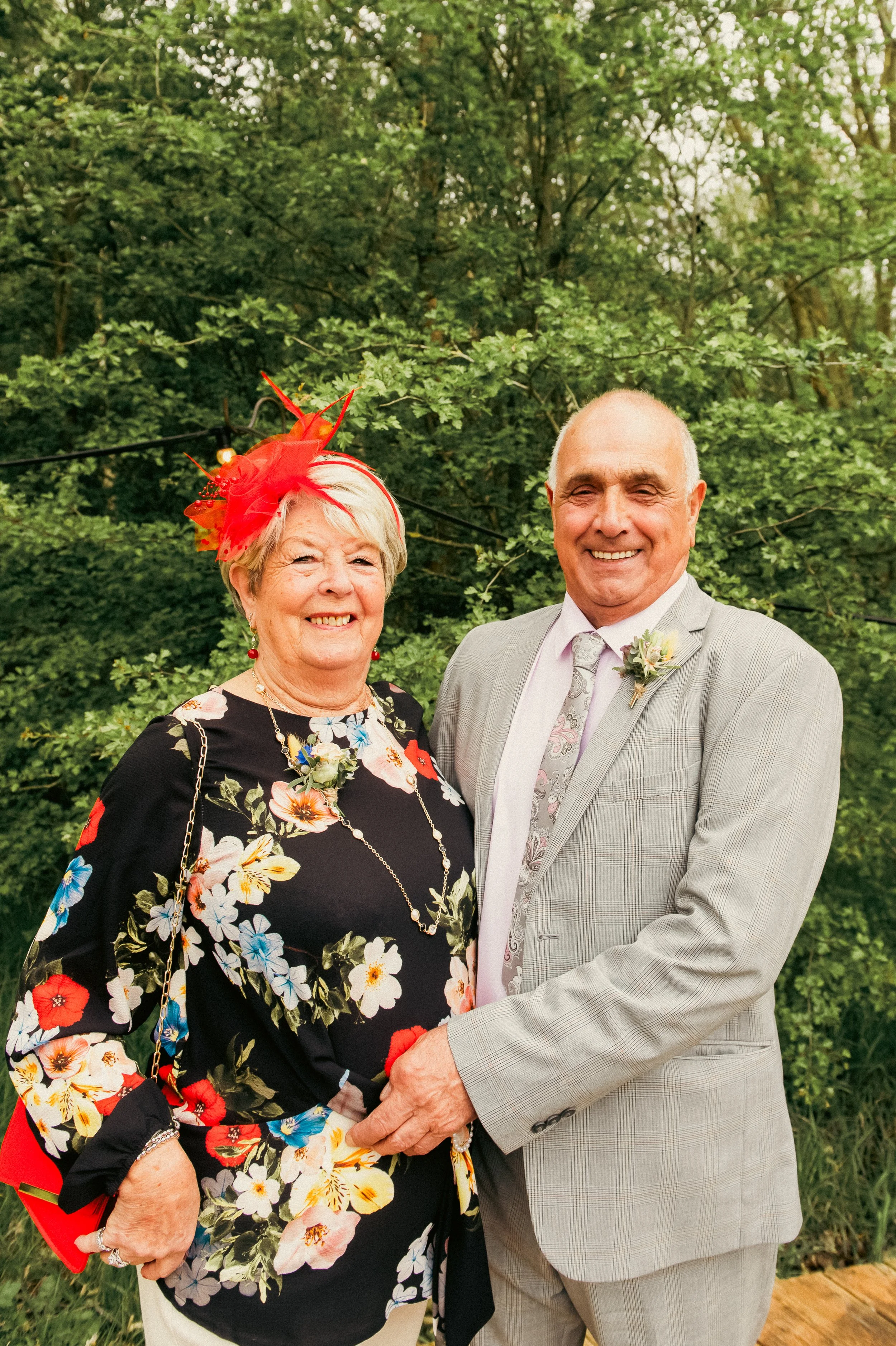 An elderly woman and man dressed in formal attire, smiling and holding hands outdoors with green trees in the background. The woman wears a black floral dress with red, yellow, white, and blue flowers, red earrings, a gold chain necklace, a red fasci