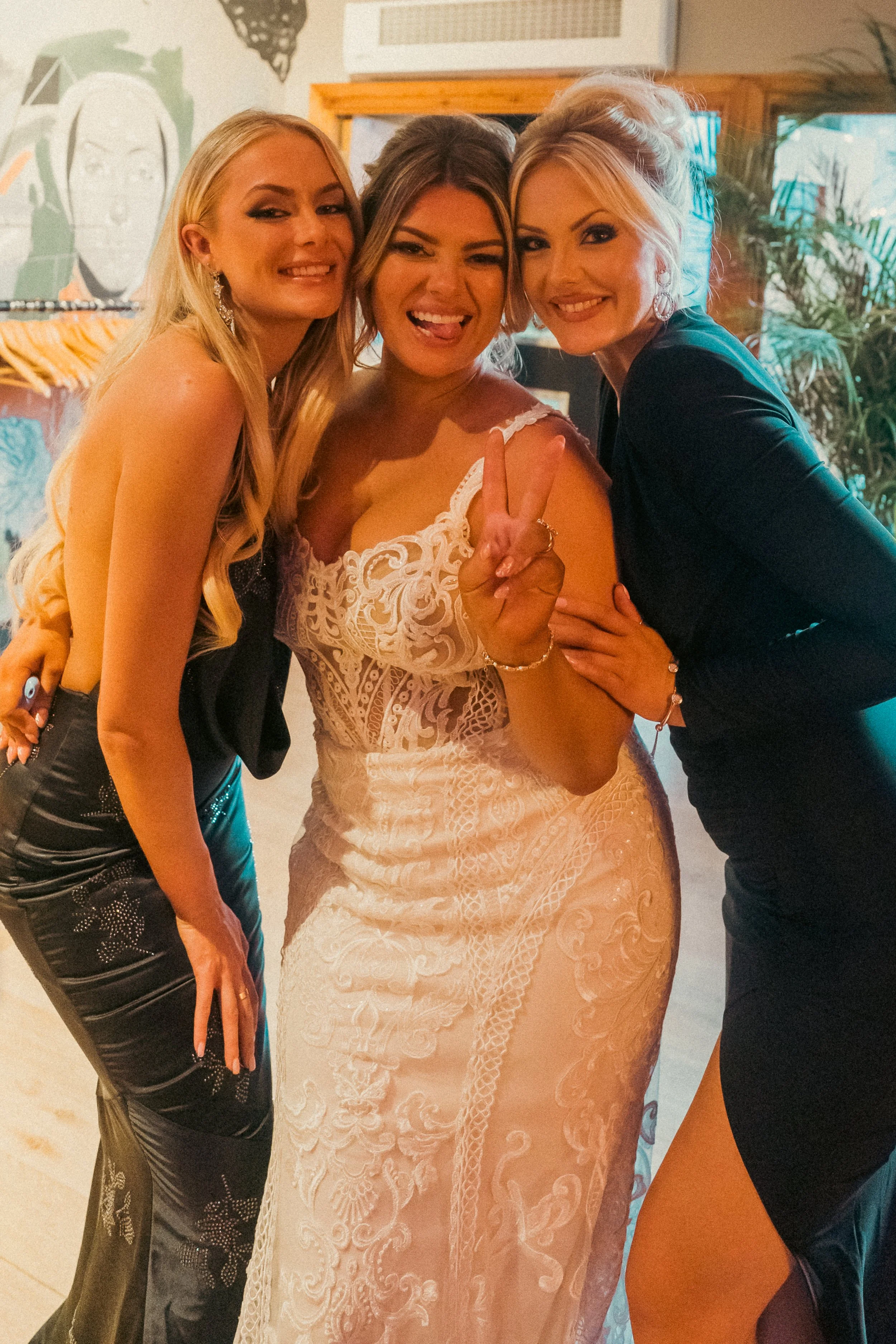 Three women smiling and posing together indoors, one woman in a white lace dress making a peace sign, and two women on either side of her.