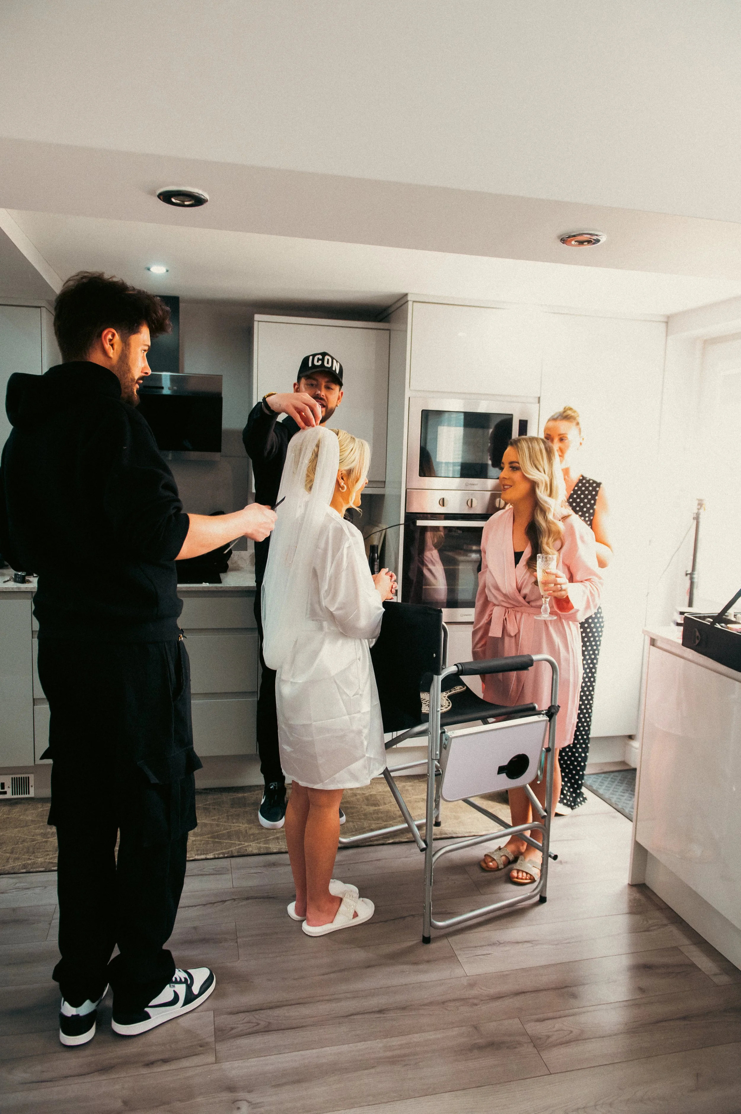 People preparing a bride in a modern kitchen, one woman in a white robe with a veil, others helping and chatting, holding drinks, with a wheelchair nearby.