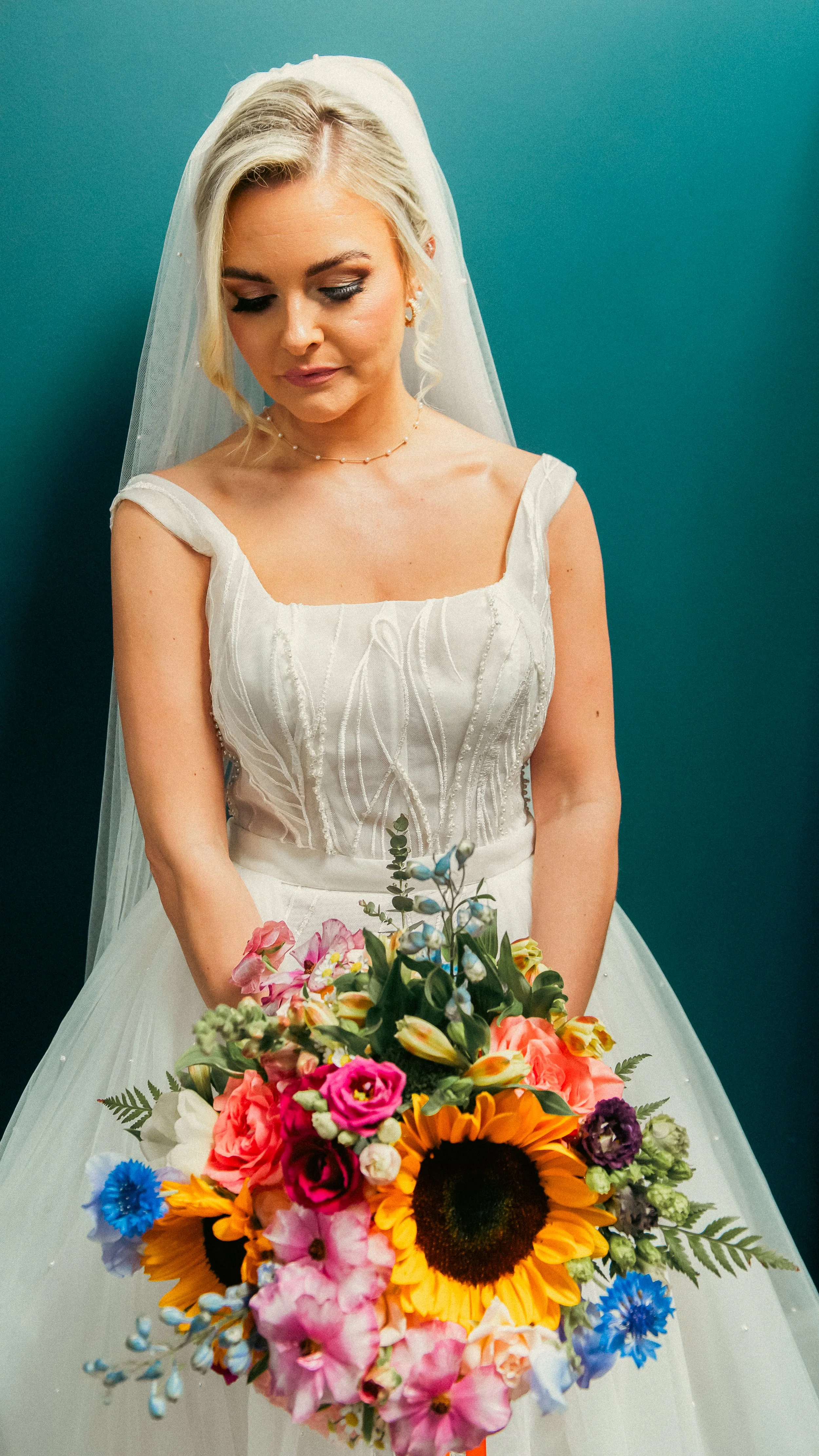 A blonde bride in a white wedding dress holding a colorful bouquet of flowers in front of a teal background.