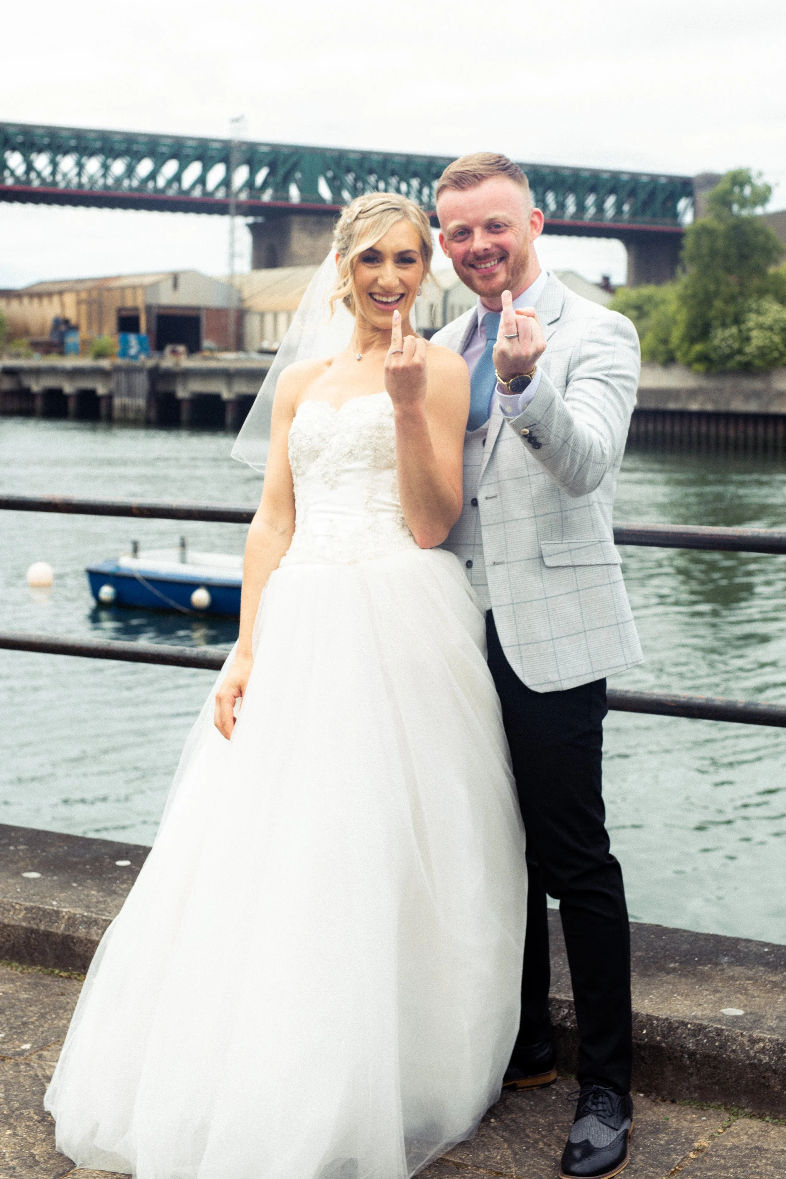 A newlywed couple standing by a waterfront, smiling, showing their middle fingers. The bride is wearing a white wedding dress and veil, and the groom is in a light gray checkered blazer, blue shirt, and black pants.