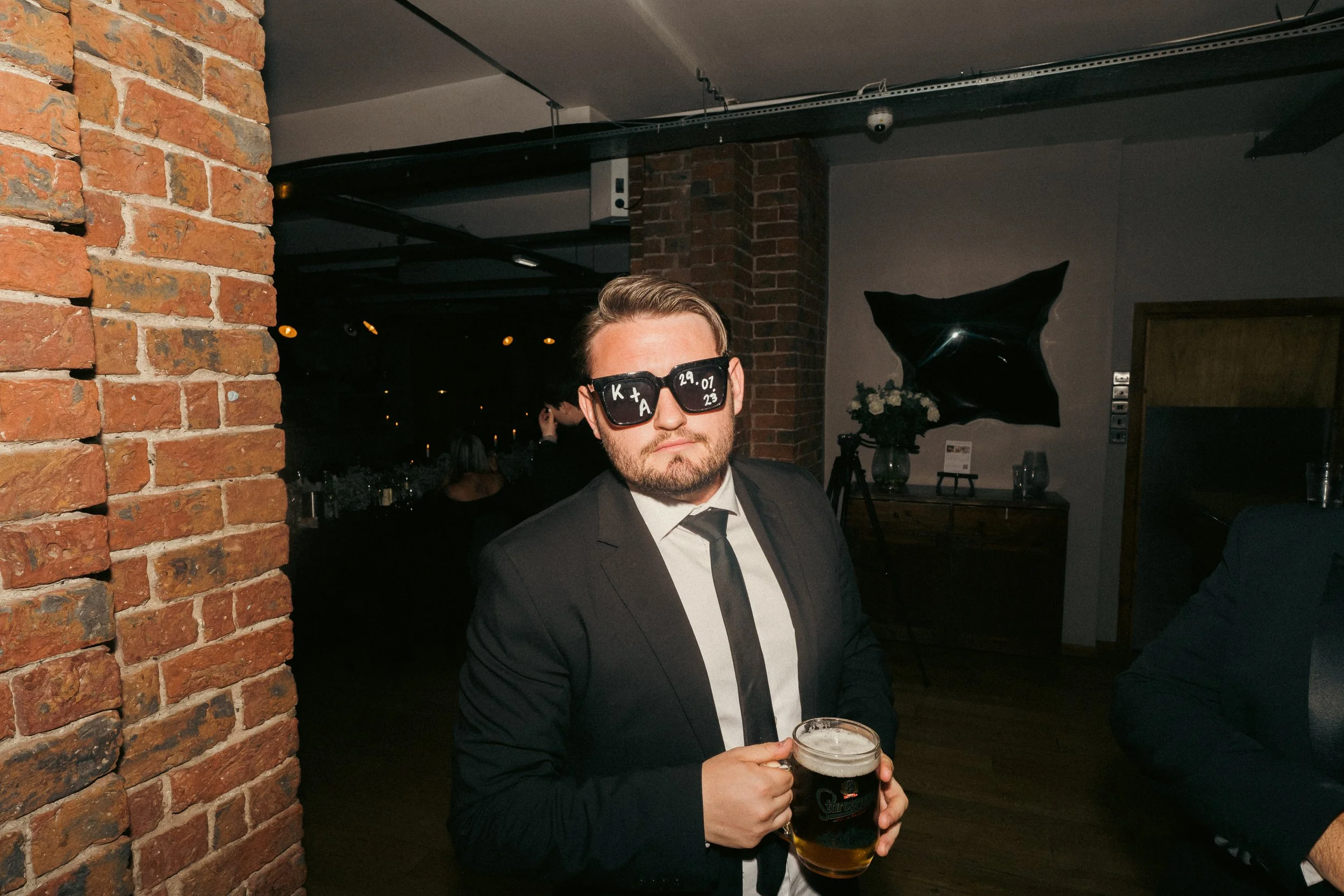 A man in a black suit and tie is holding a glass of dark beer, wearing sunglasses with handwritten initials and date, standing in a dimly lit bar or restaurant with exposed brick walls.
