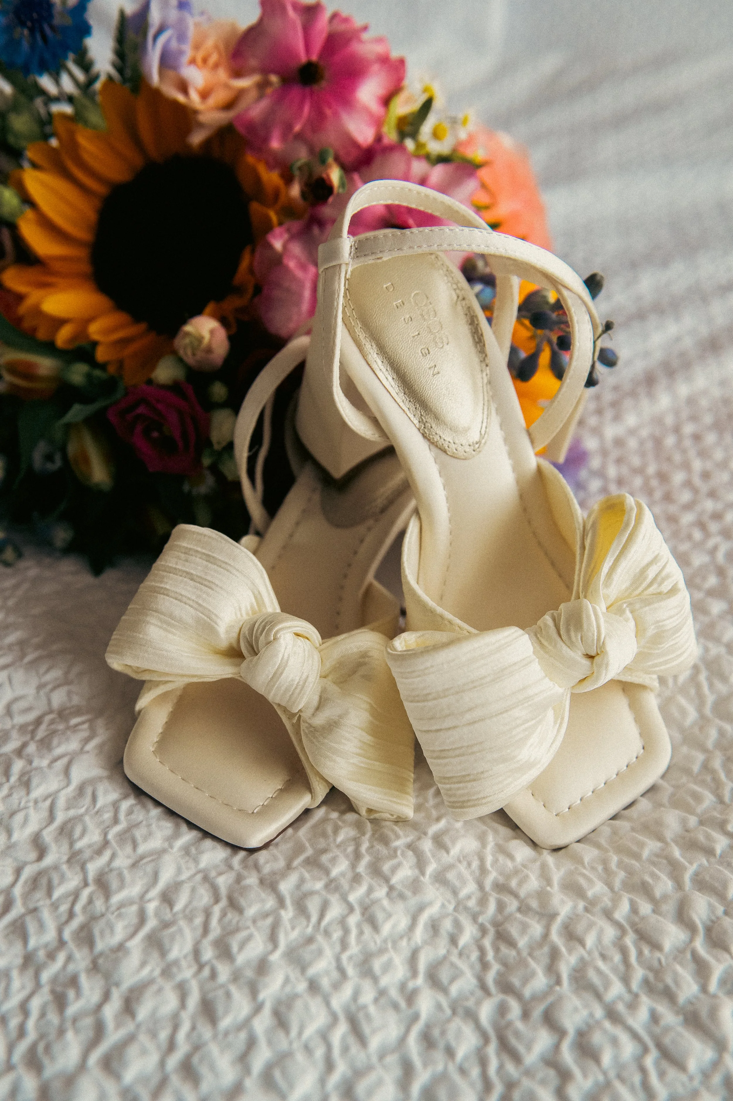 Cream-colored high-heeled shoes with large fabric bows on the front, placed on a textured white surface with a colorful bouquet of flowers in the background.