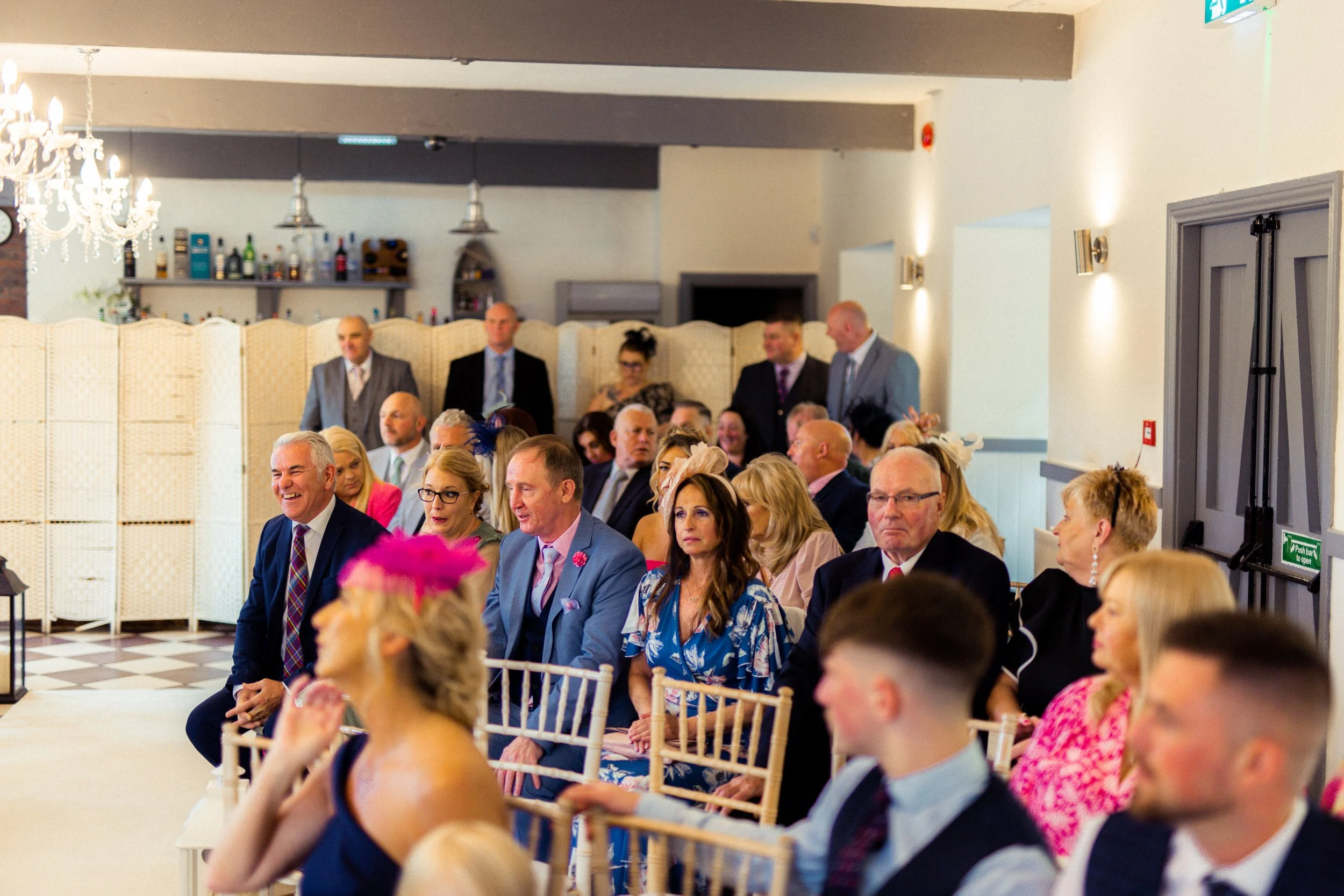 Guests seated in rows at a wedding ceremony, dressed in formal attire, with some wearing fascinators and hats, in a decorated indoor venue with chandelier and bar in background.