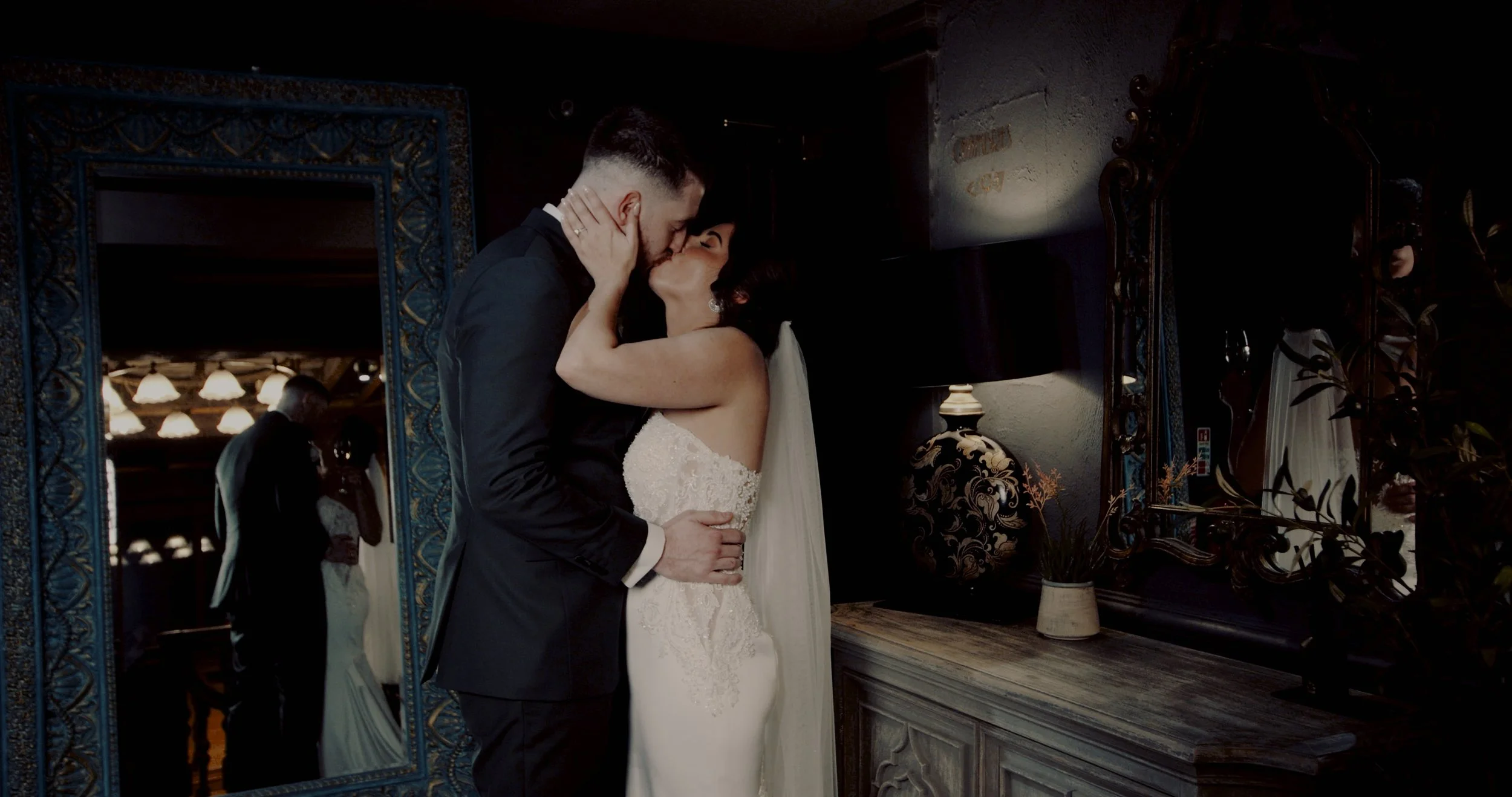 A bride and groom sharing a kiss during their wedding in a dimly lit room, with a reflection of their embrace in a tall ornate mirror.