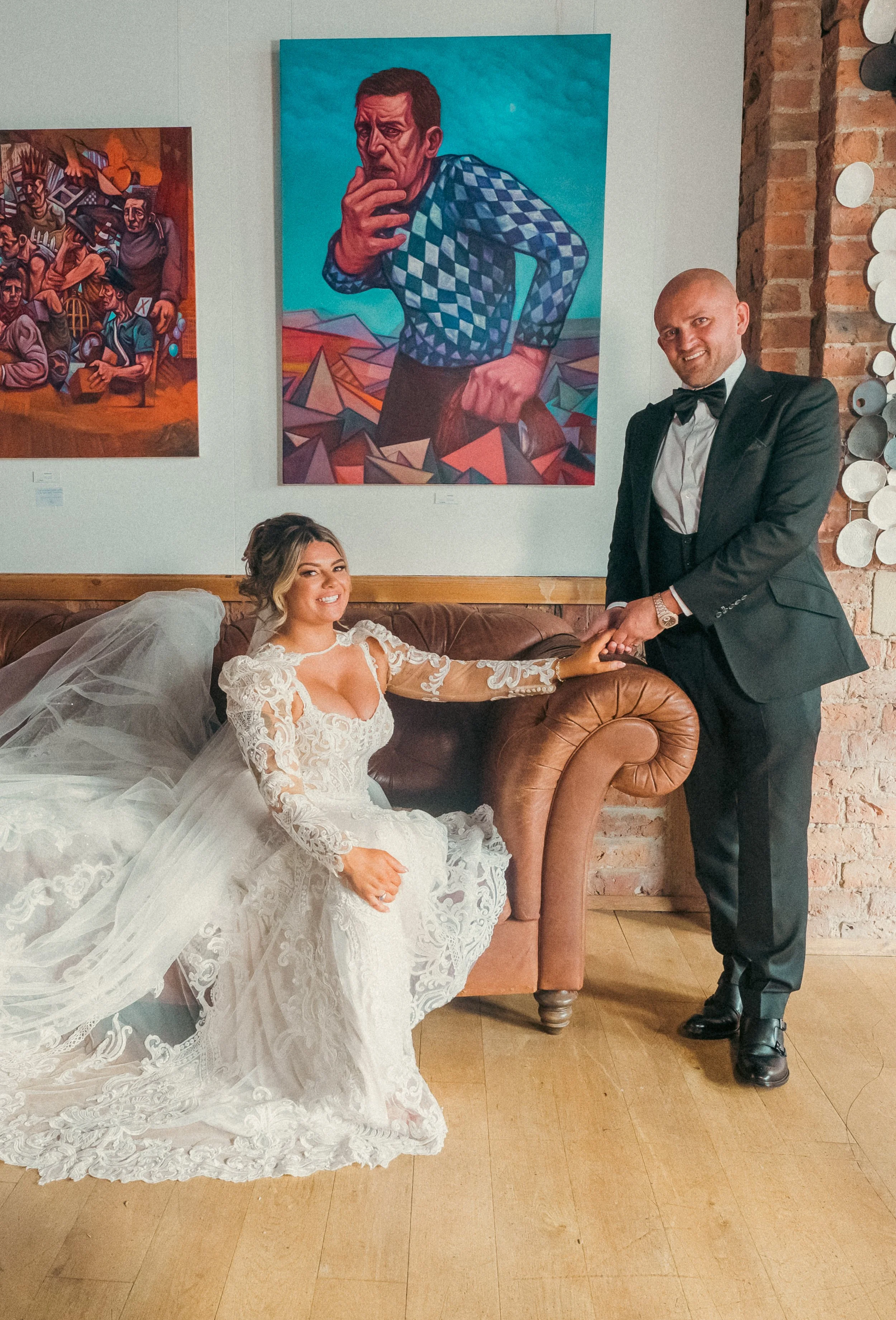 A bride in a white lace wedding gown sitting on a leather sofa, holding hands with a groom in a black tuxedo, standing beside her. They are in an art gallery with colorful paintings on the wall behind them, including a large portrait of a man in a ch