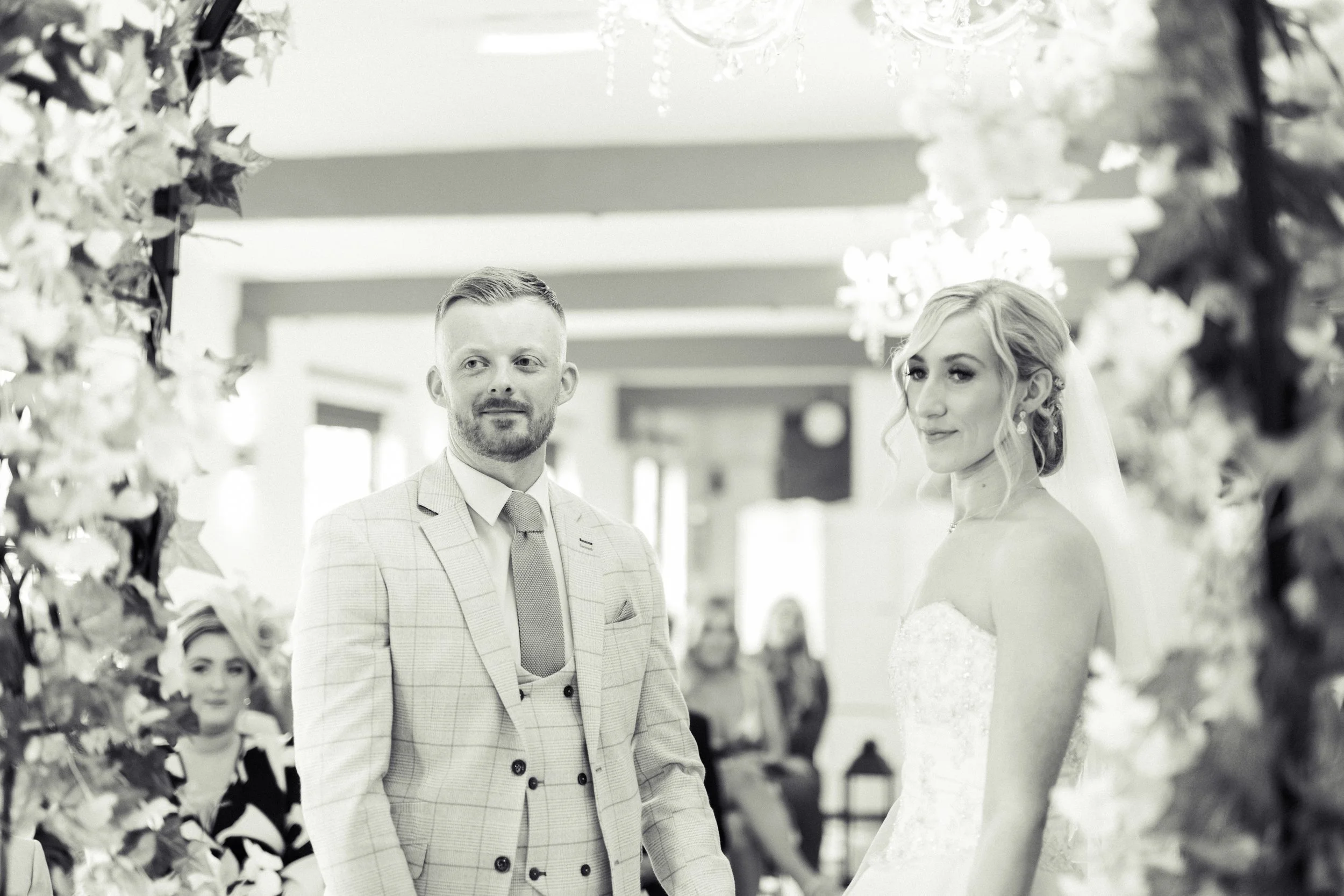 A couple standing during a wedding ceremony, with the bride on the right wearing a white wedding dress and veil, and the groom on the left in a light-colored suit, indoors decorated with flowers and chandeliers.