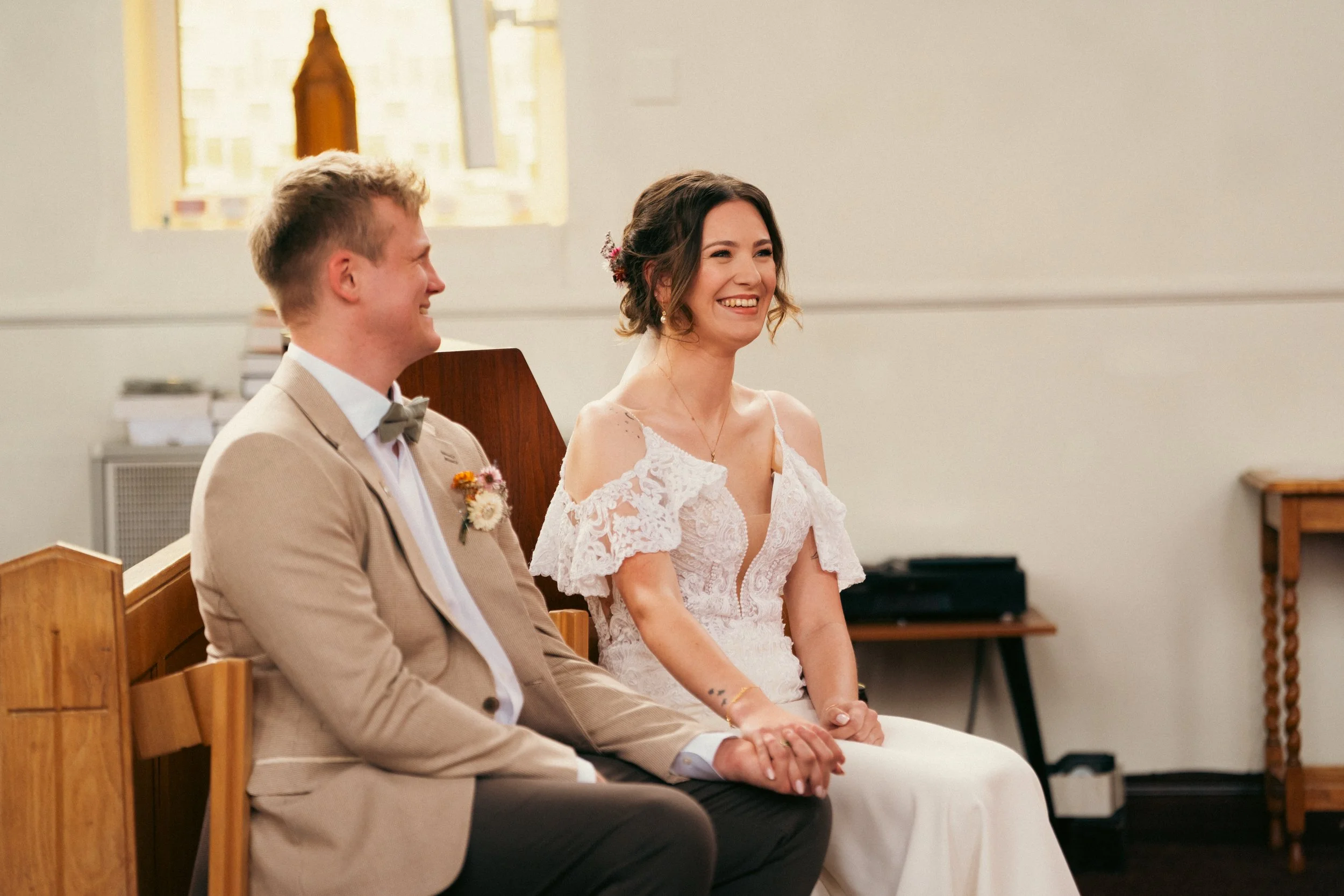 A smiling woman in a white lace off-shoulder wedding dress and a man in a tan blazer with a boutonniere sit together holding hands inside a church during a wedding ceremony.