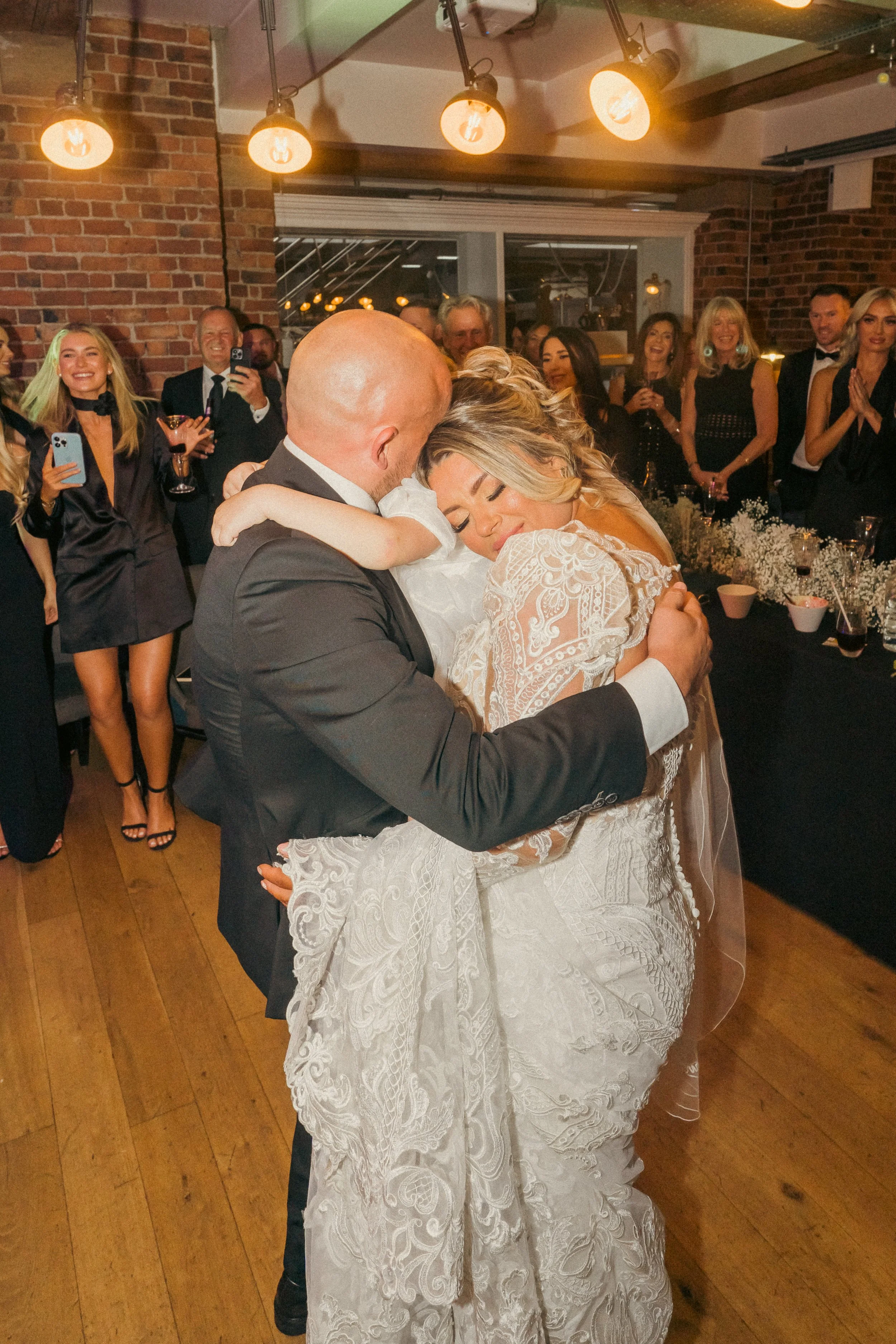 A bride and groom share an embrace during their wedding reception, surrounded by happy guests in a warmly lit, rustic venue.
