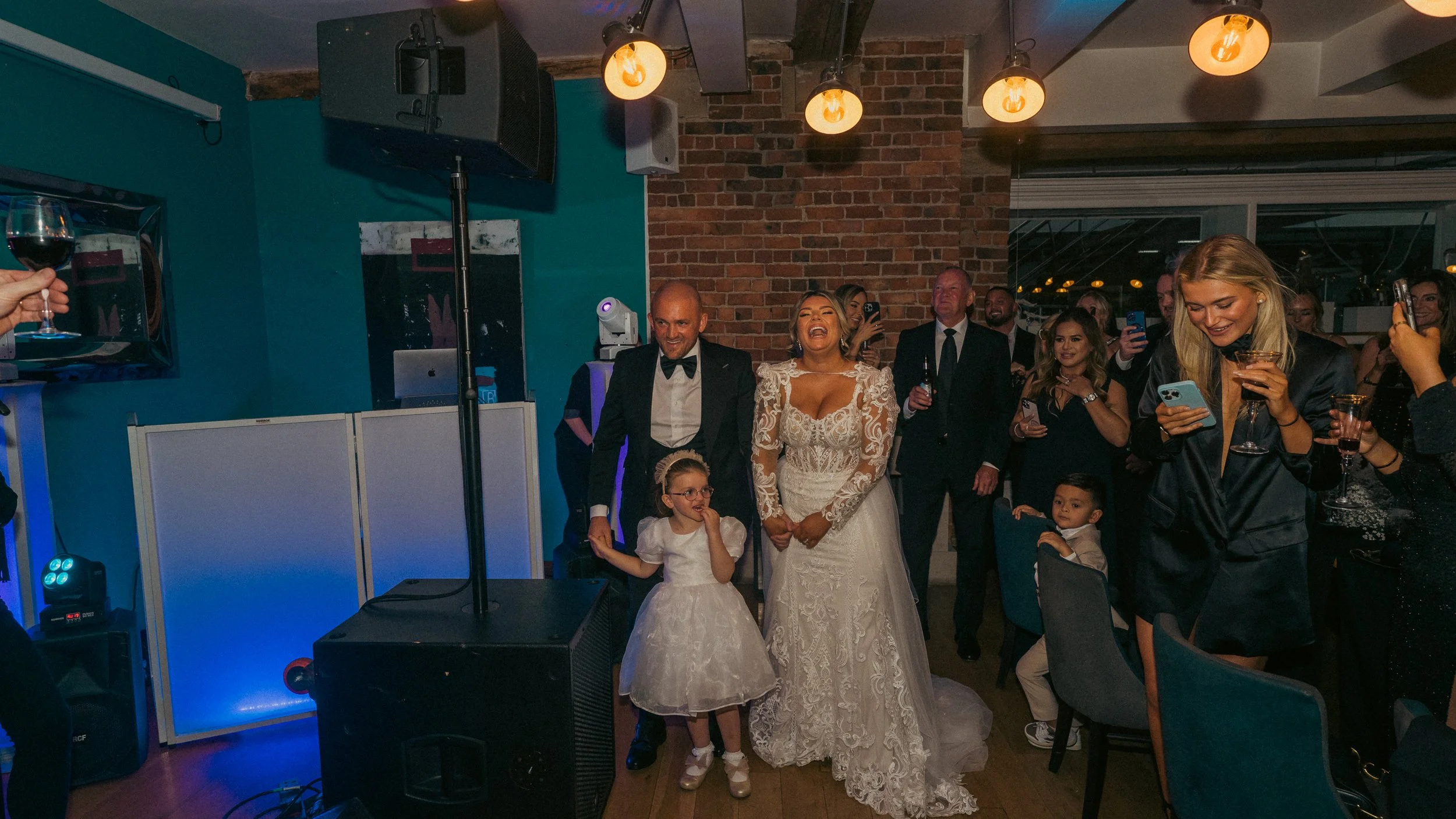 A wedding reception with a bride in a lace wedding gown, a groom in a tuxedo, and children, surrounded by guests taking photos and enjoying drinks in a decorated indoor venue with brick wall and warm lighting.