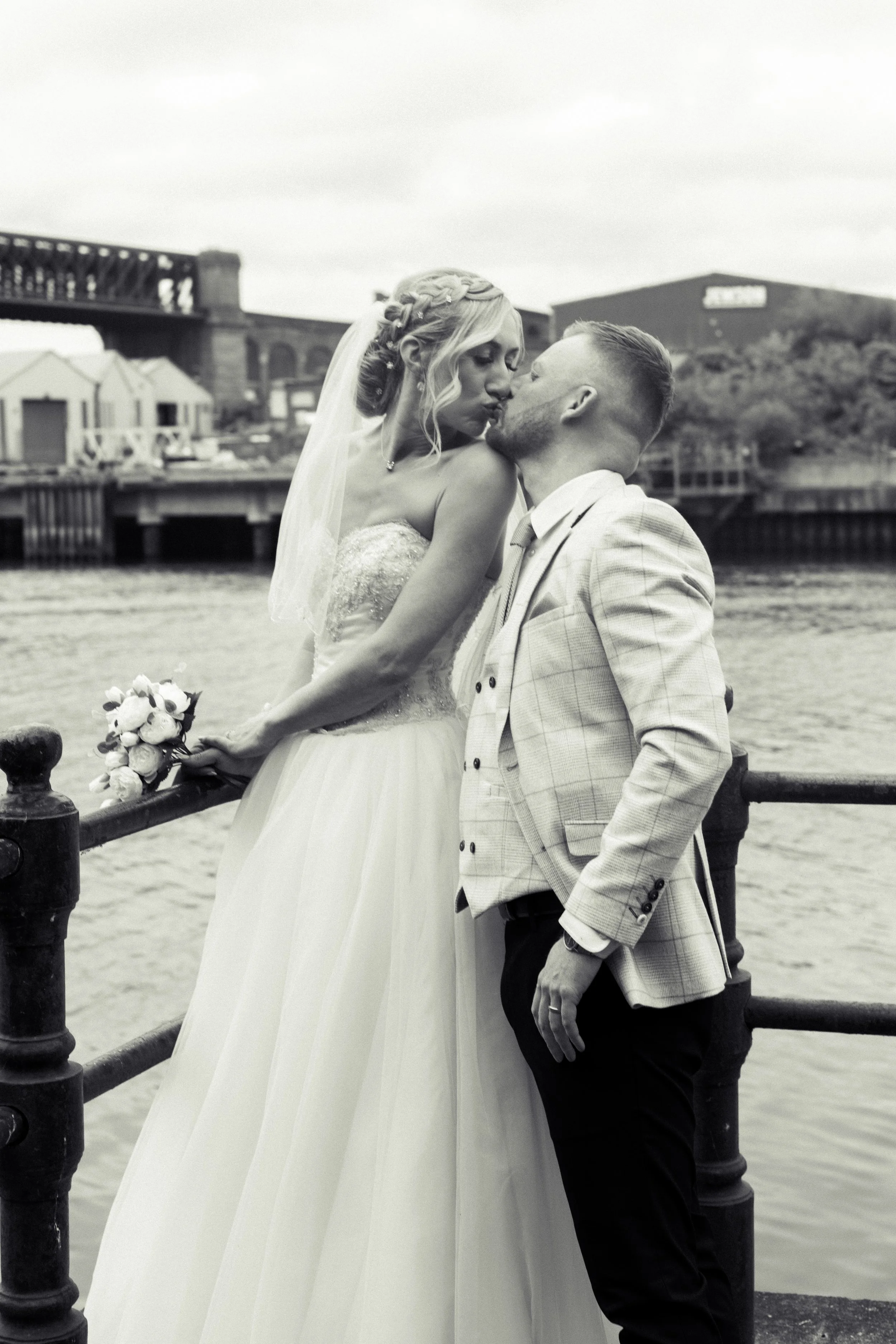 A couple in wedding attire sharing a kiss by the water, with a bridge and buildings in the background.