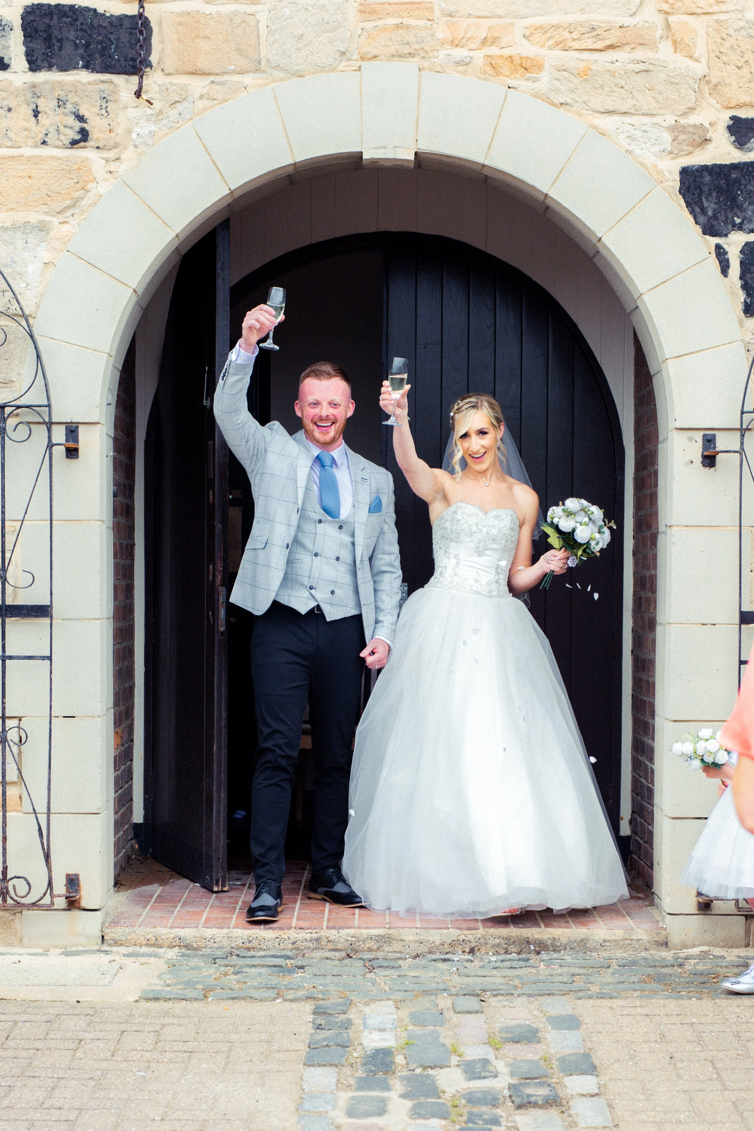 A newly married couple celebrating outside a stone archway, holding champagne glasses. The groom is dressed in a light gray checked tuxedo with a blue tie, and the bride is wearing a white wedding gown and holding a bouquet of white flowers.