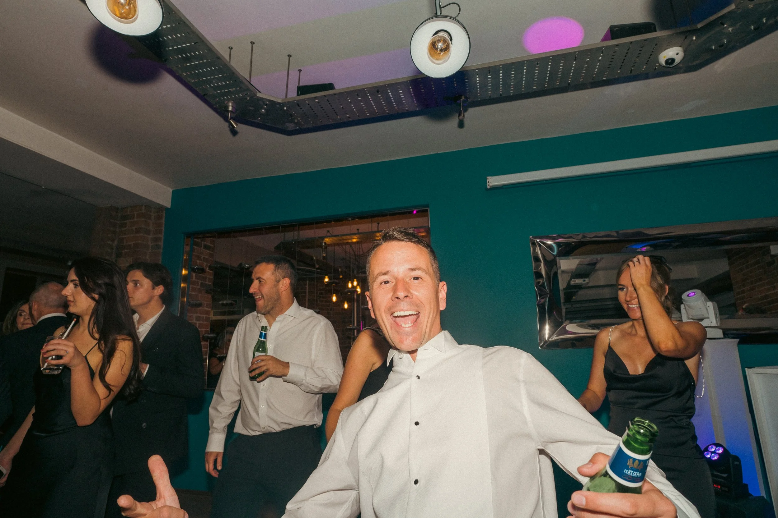 People socializing at a party, with a man smiling in the foreground holding a beer bottle, others drinking and chatting in the background.