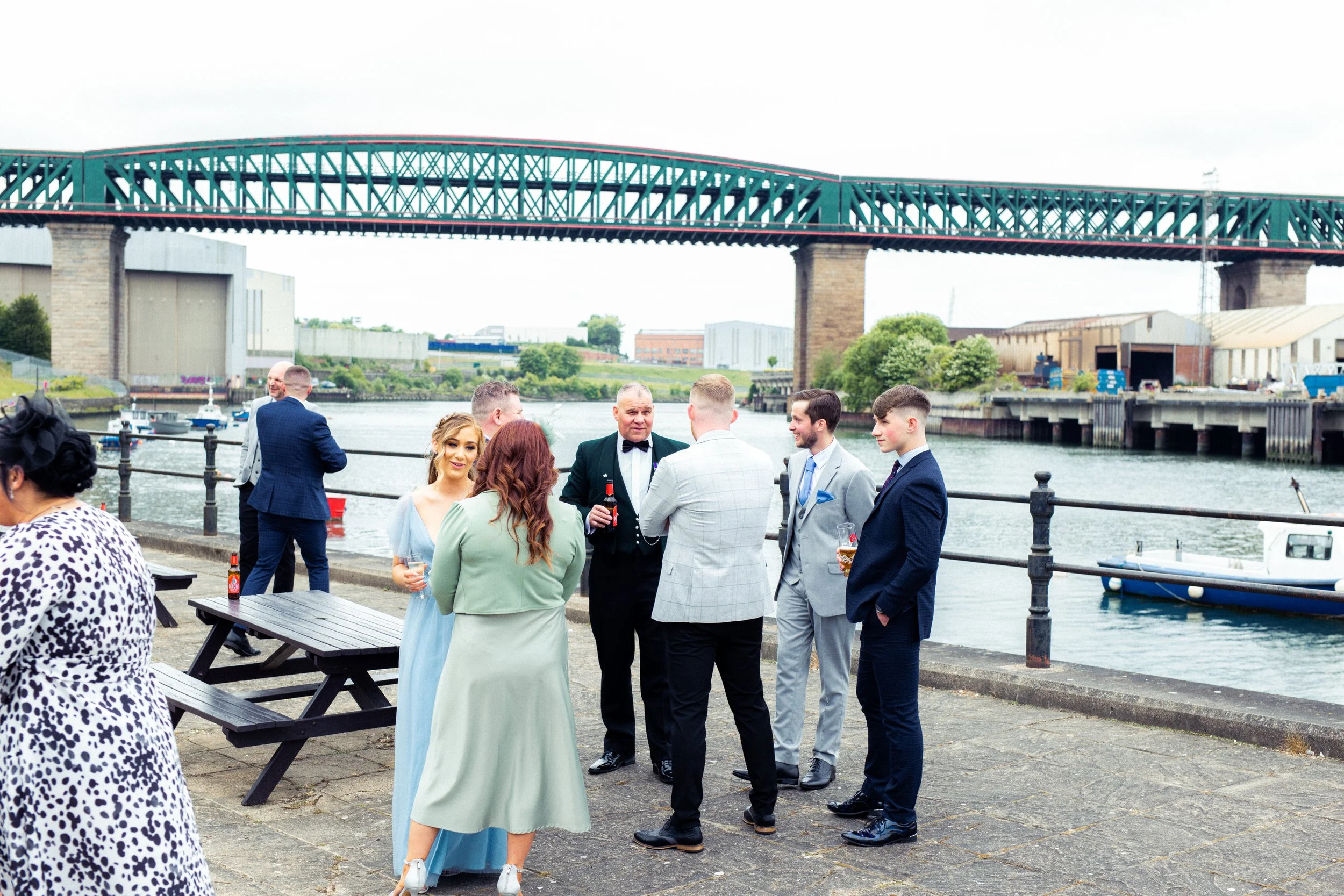 A group of people dressed in formal attire socializing near a waterfront with a bridge in the background, some holding drinks.