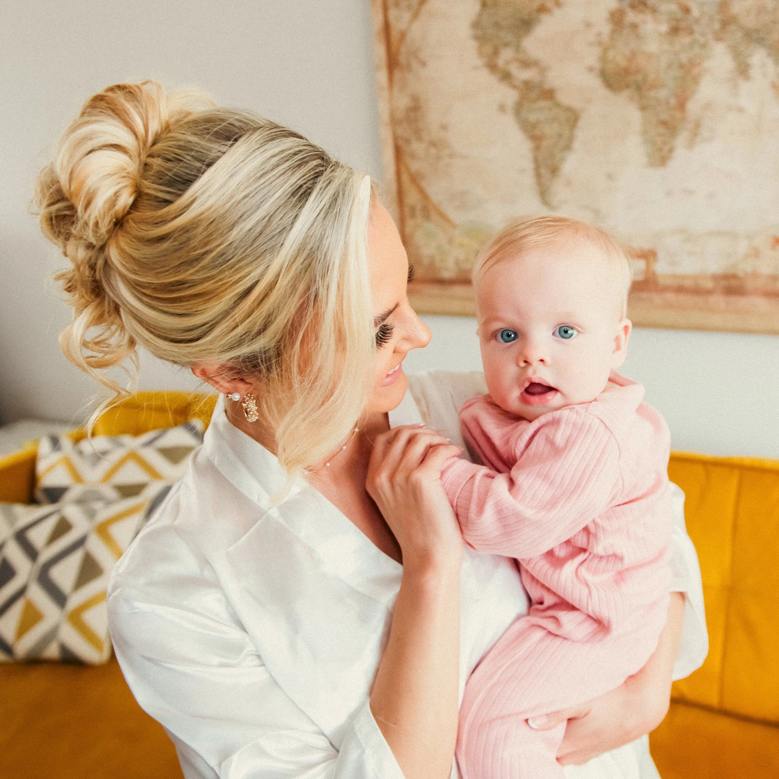 A blonde woman with styled hair holding a baby girl with blue eyes wearing a pink outfit in a living room with a world map on the wall and a yellow couch.