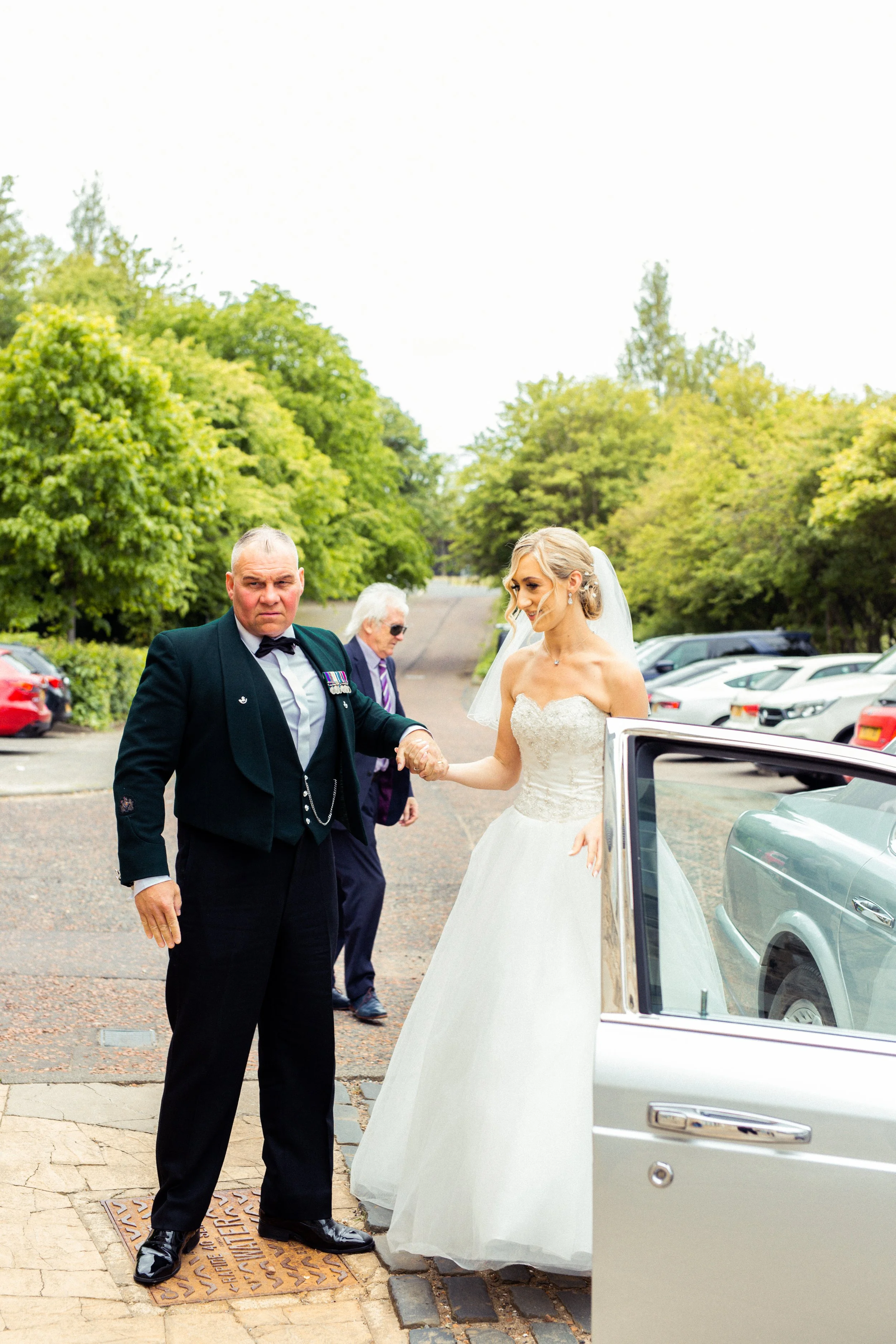 A bride in a white wedding gown getting out of a car while holding hands with an older man in a military uniform during a wedding ceremony.