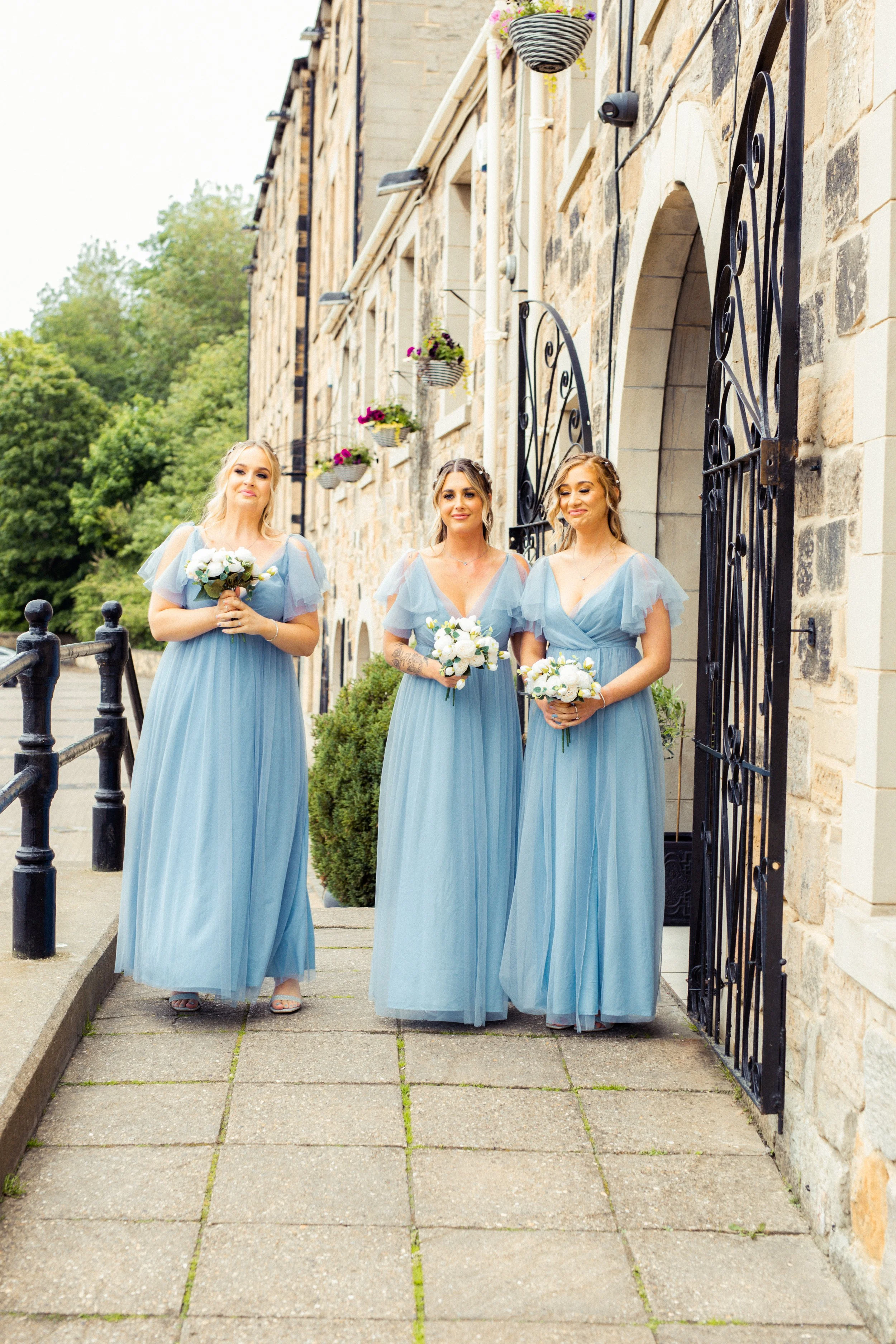 Three women in light blue bridesmaid dresses holding bouquets, standing on a sidewalk in front of a stone building with black wrought iron gates and hanging flower baskets during daylight.