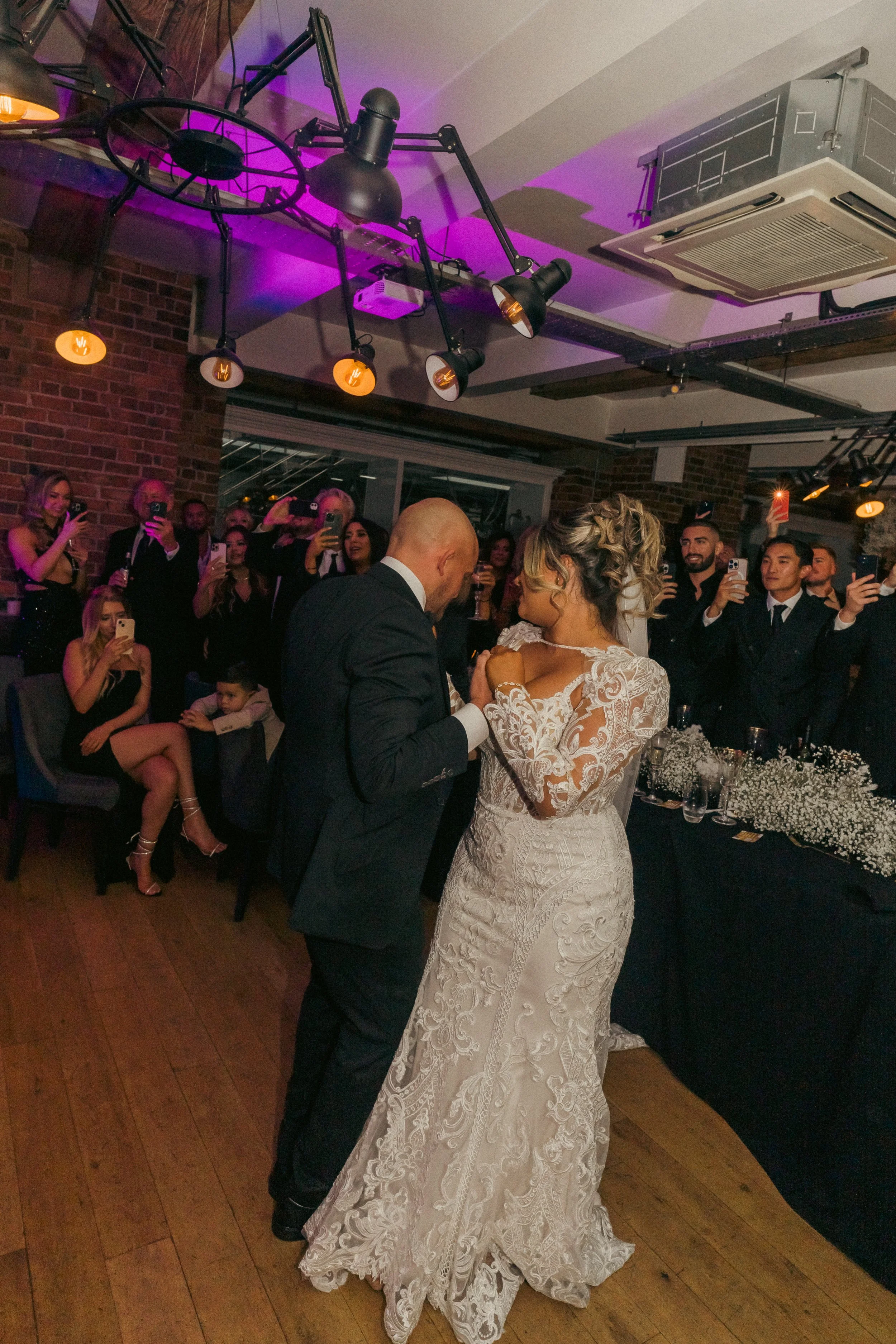 A bride and groom dancing at their wedding reception surrounded by guests taking photos.