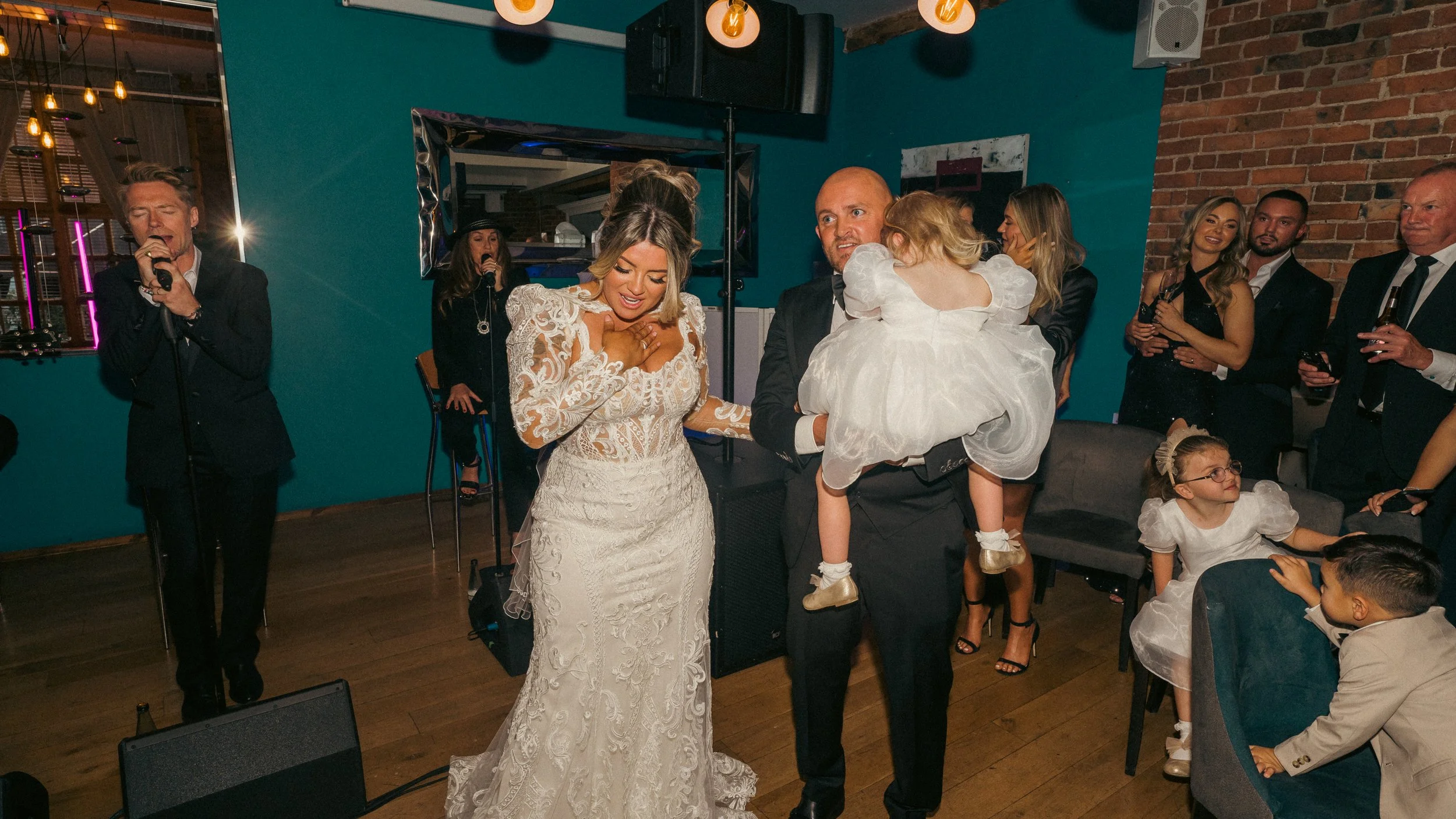 At a wedding reception, a woman in a white wedding dress and a man in a black tuxedo are holding a young girl in a white dress. Several guests are watching, some smiling and taking photos. A singer is performing with a microphone on the left.