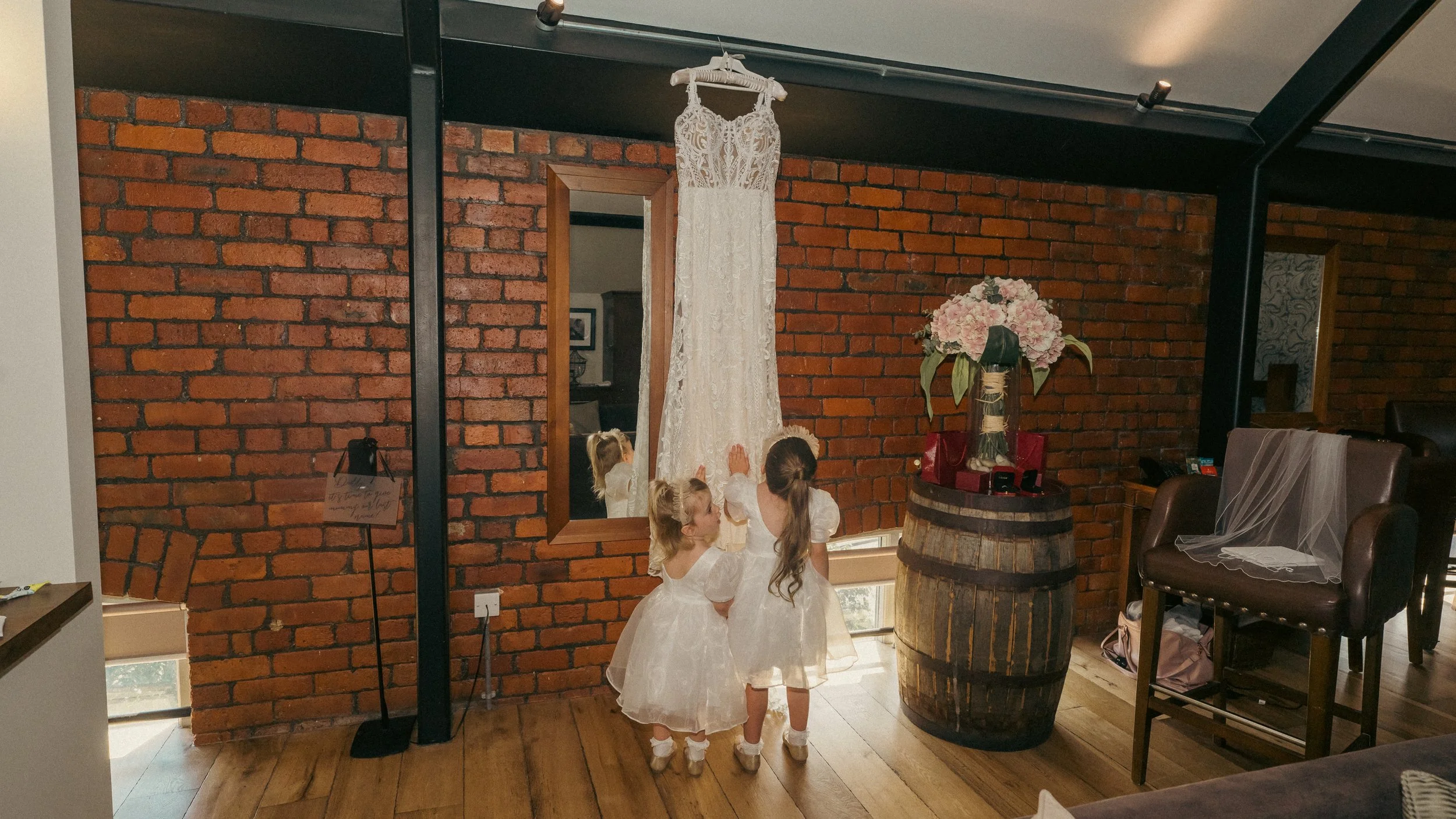 Three young girls in white dresses, likely flower girls, touching a hanging wedding dress inside a room with exposed brick walls and wooden floors.