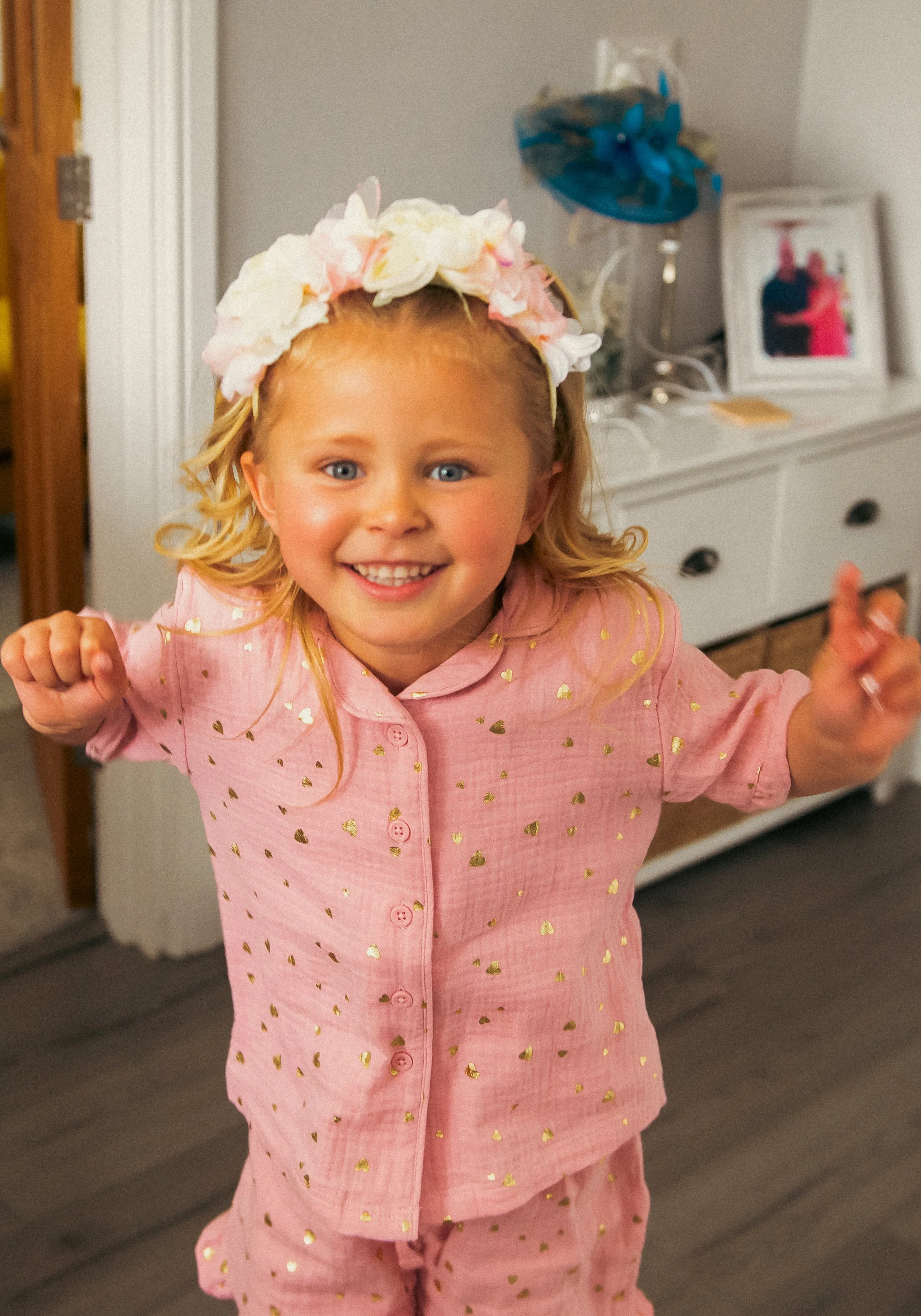 A smiling young girl wearing pink pajamas decorated with gold hearts and a floral headband, celebrating indoors.