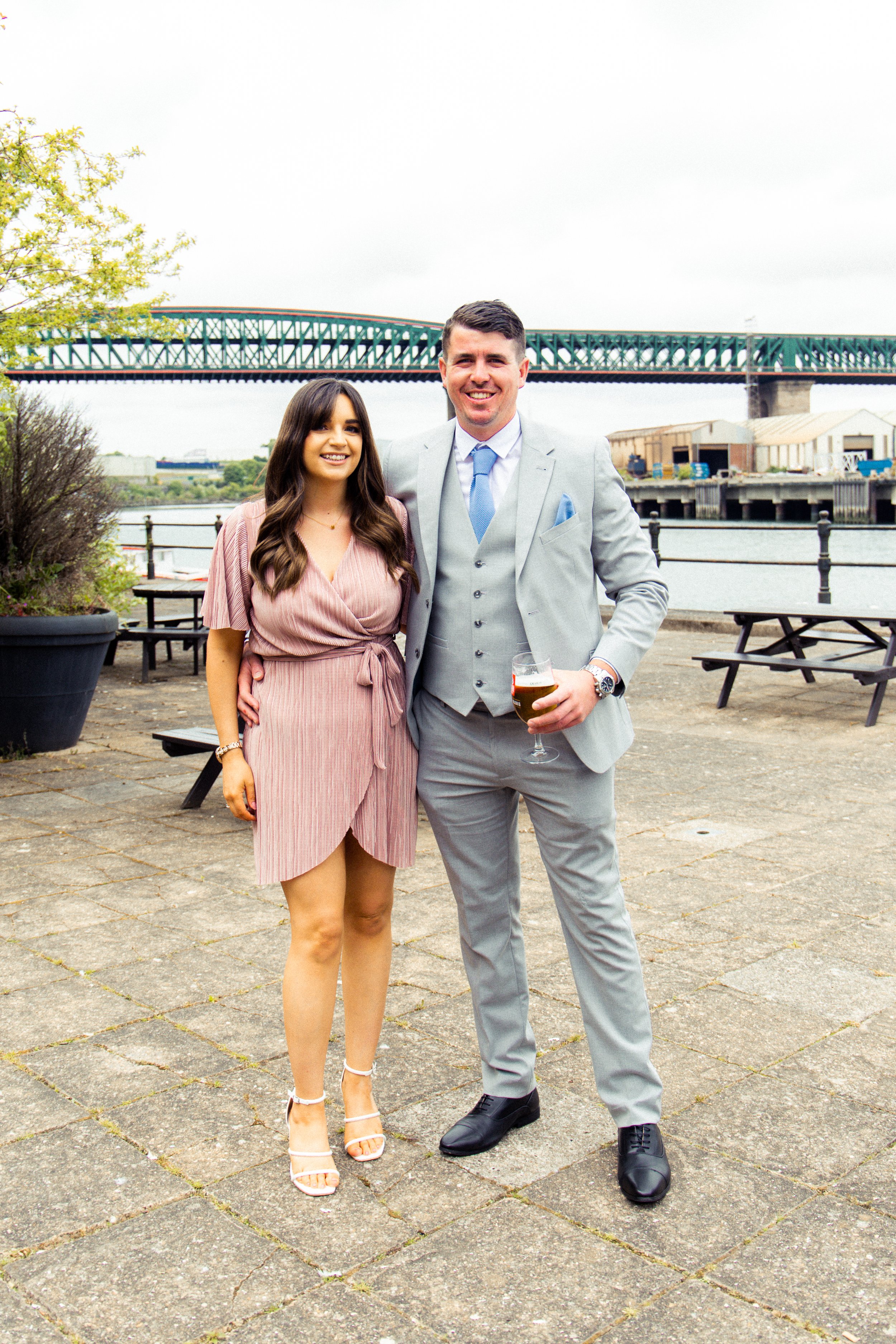 A man and a woman standing outdoors by a waterfront, smiling at the camera. The woman is wearing a pink dress and heeled sandals, and the man is in a light gray suit holding a glass of beer.