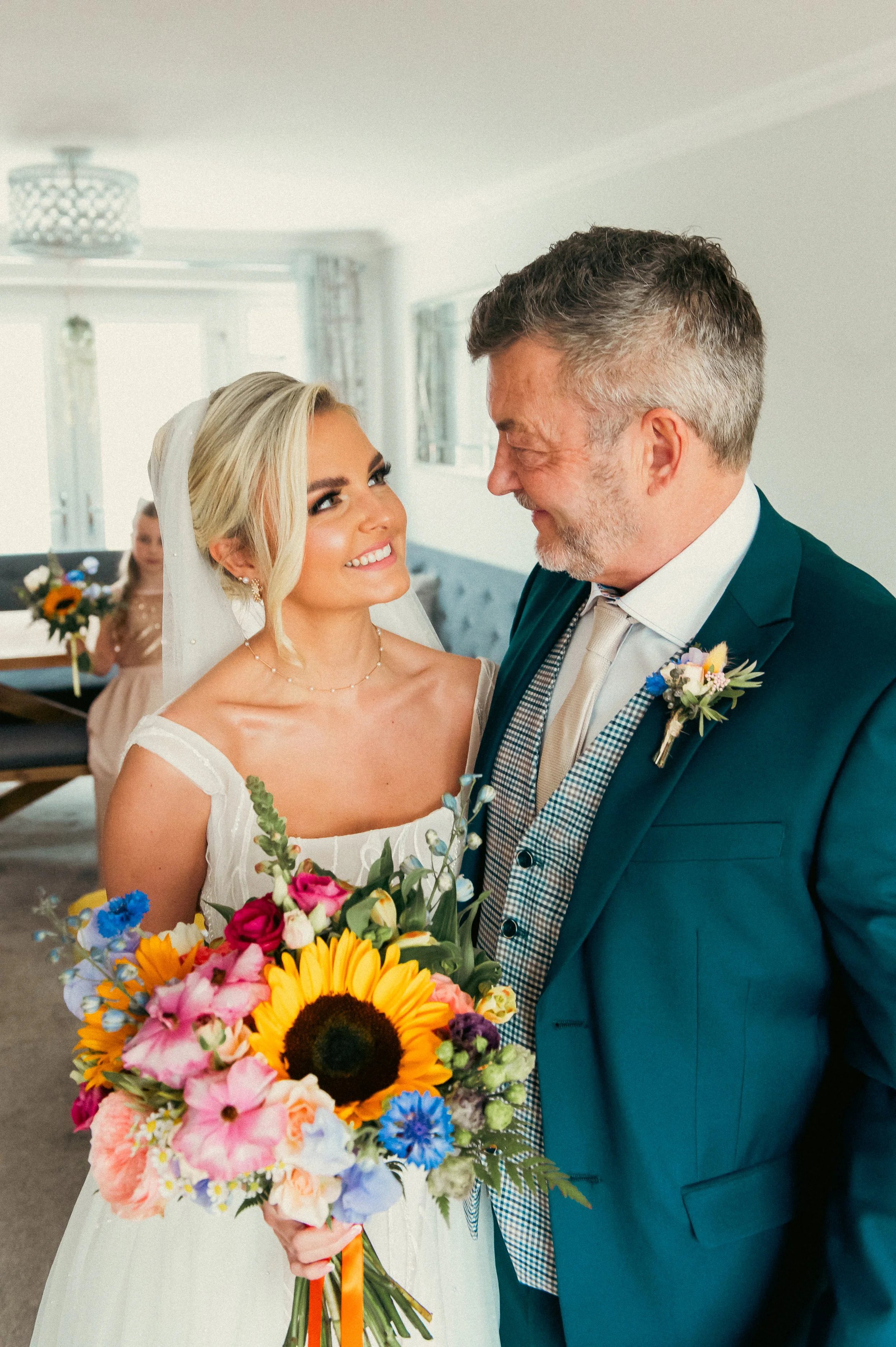 A bride holding a colorful bouquet smiling at an older man in a teal suit, likely her father, at a wedding celebration indoors.