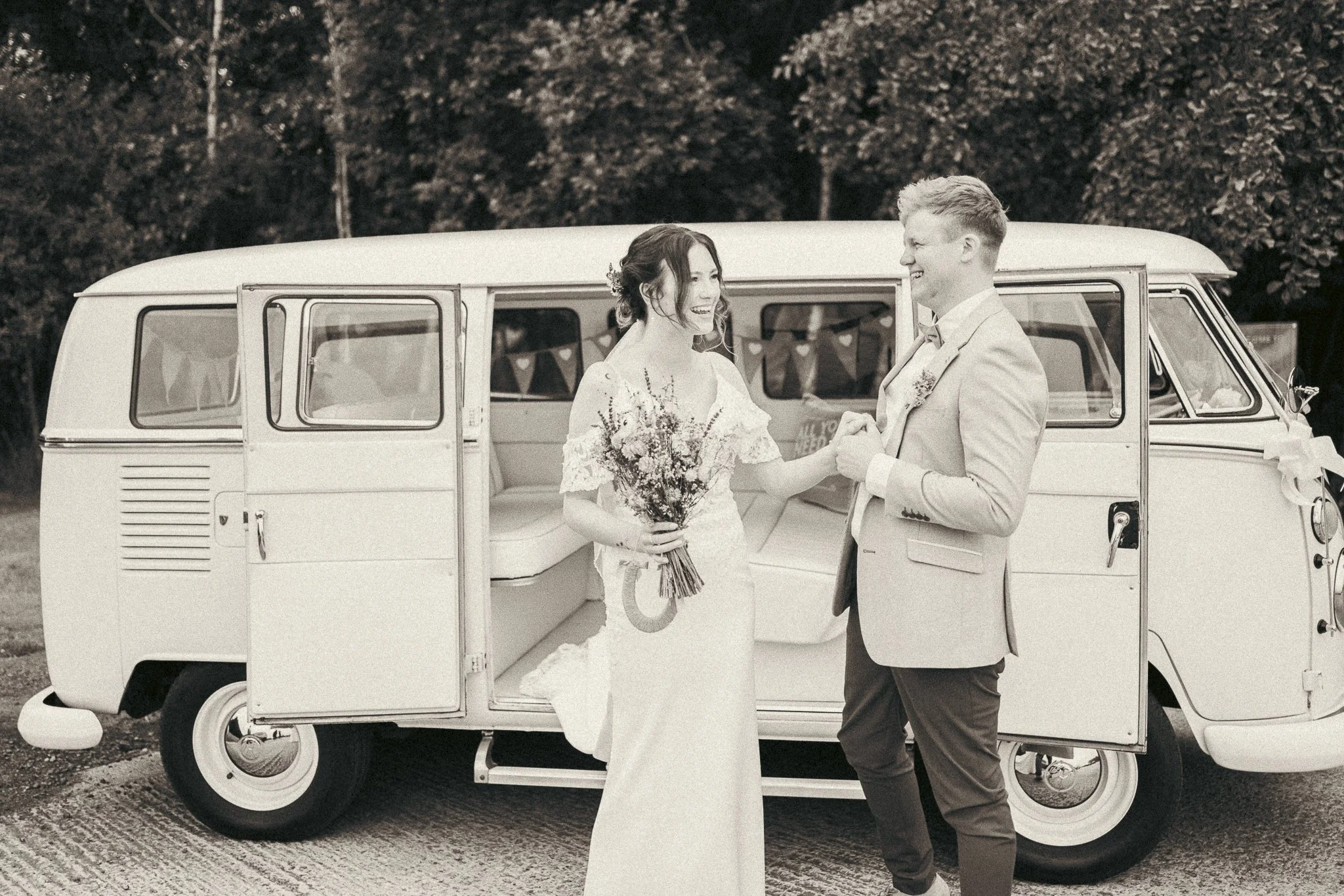 A bride and groom exchanging vows in front of a vintage camper van, with the bride holding a bouquet and the groom wearing a light-colored suit, outdoors with trees in the background.