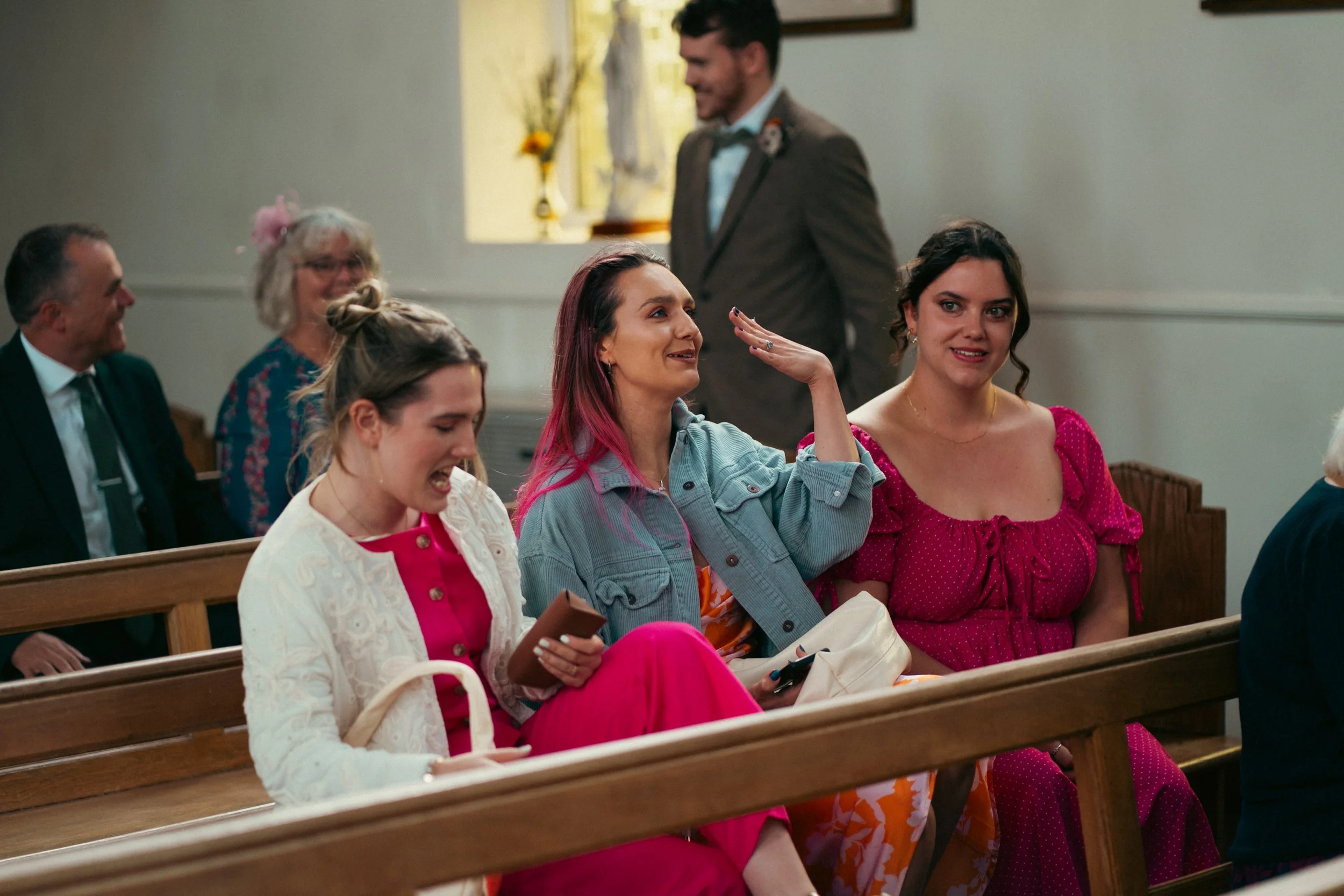 People sitting in church pews, attending a wedding, smiling, and engaging in conversation.