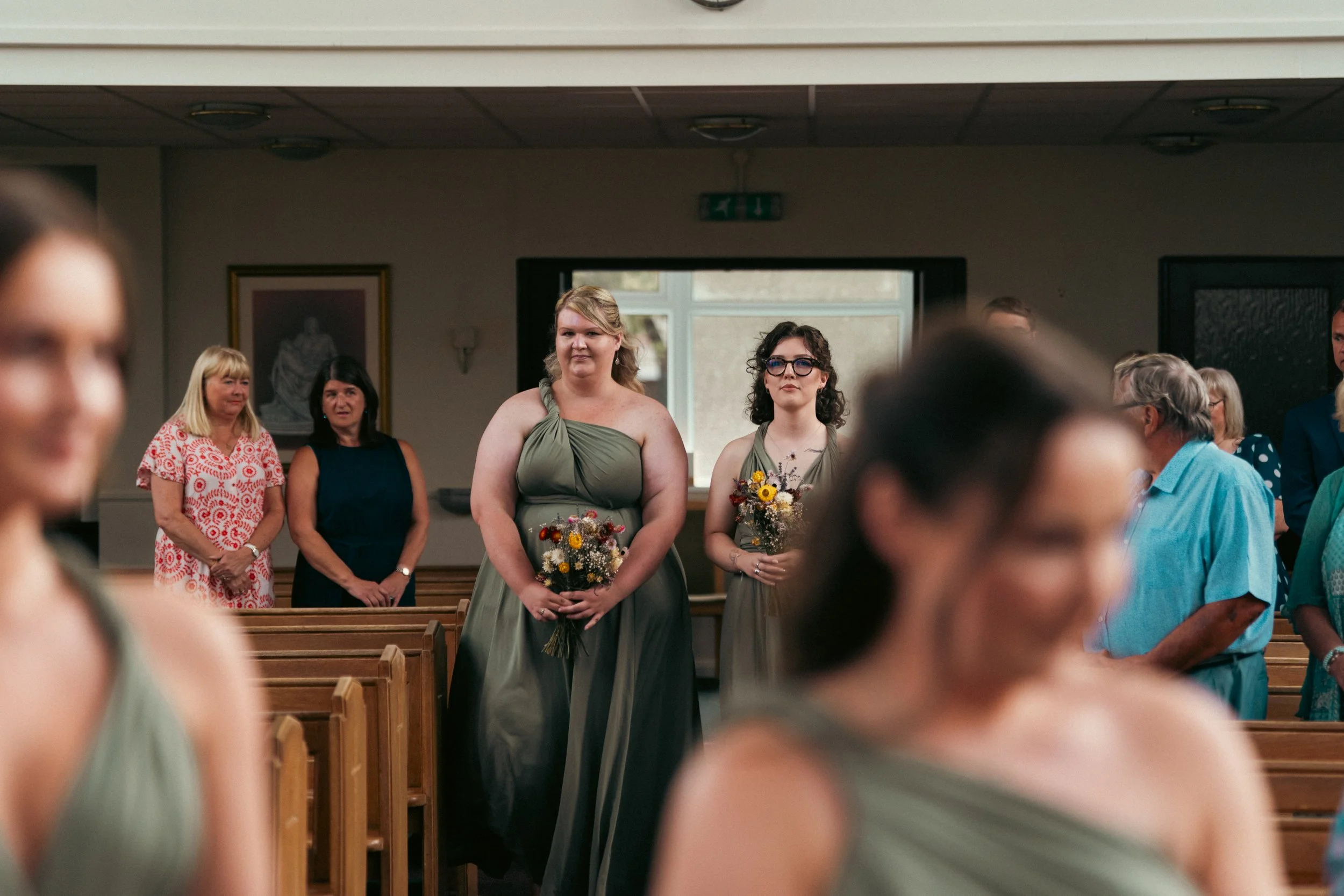 Women in formal dresses holding bouquets standing in church during a wedding ceremony with seated guests around them.