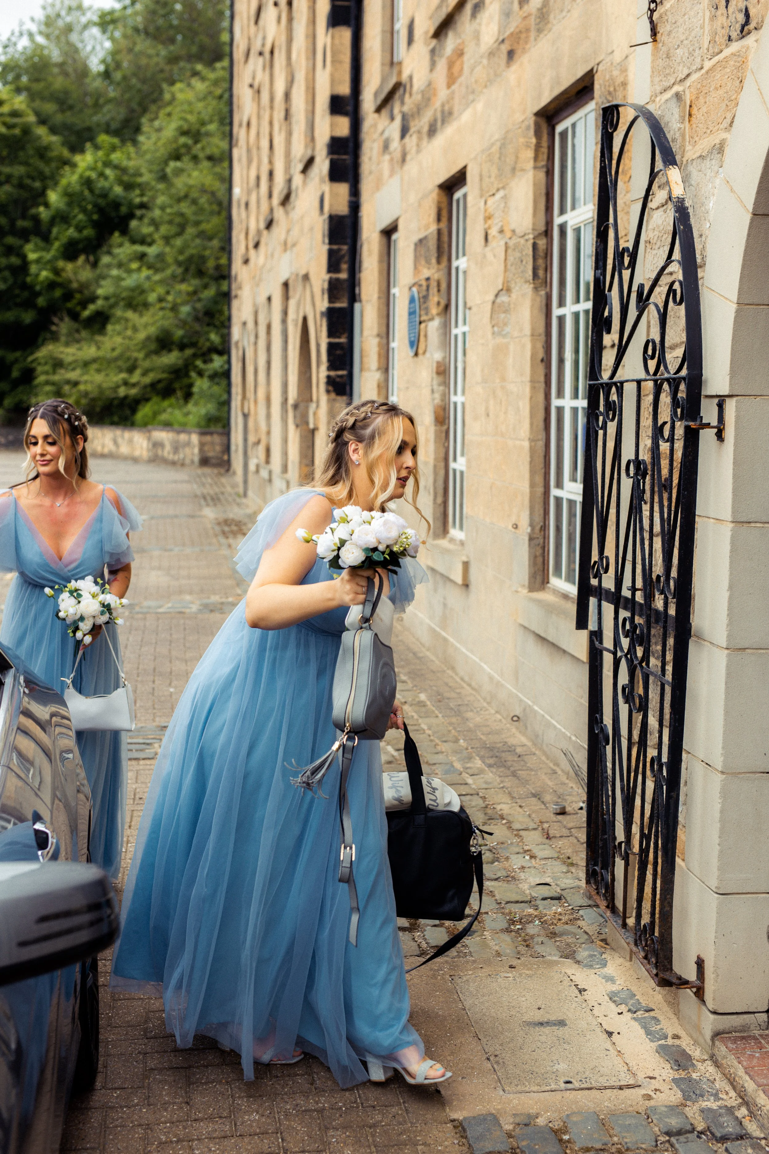 Two women in blue dresses with flower bouquets standing outside near an iron gate on a cobblestone street.