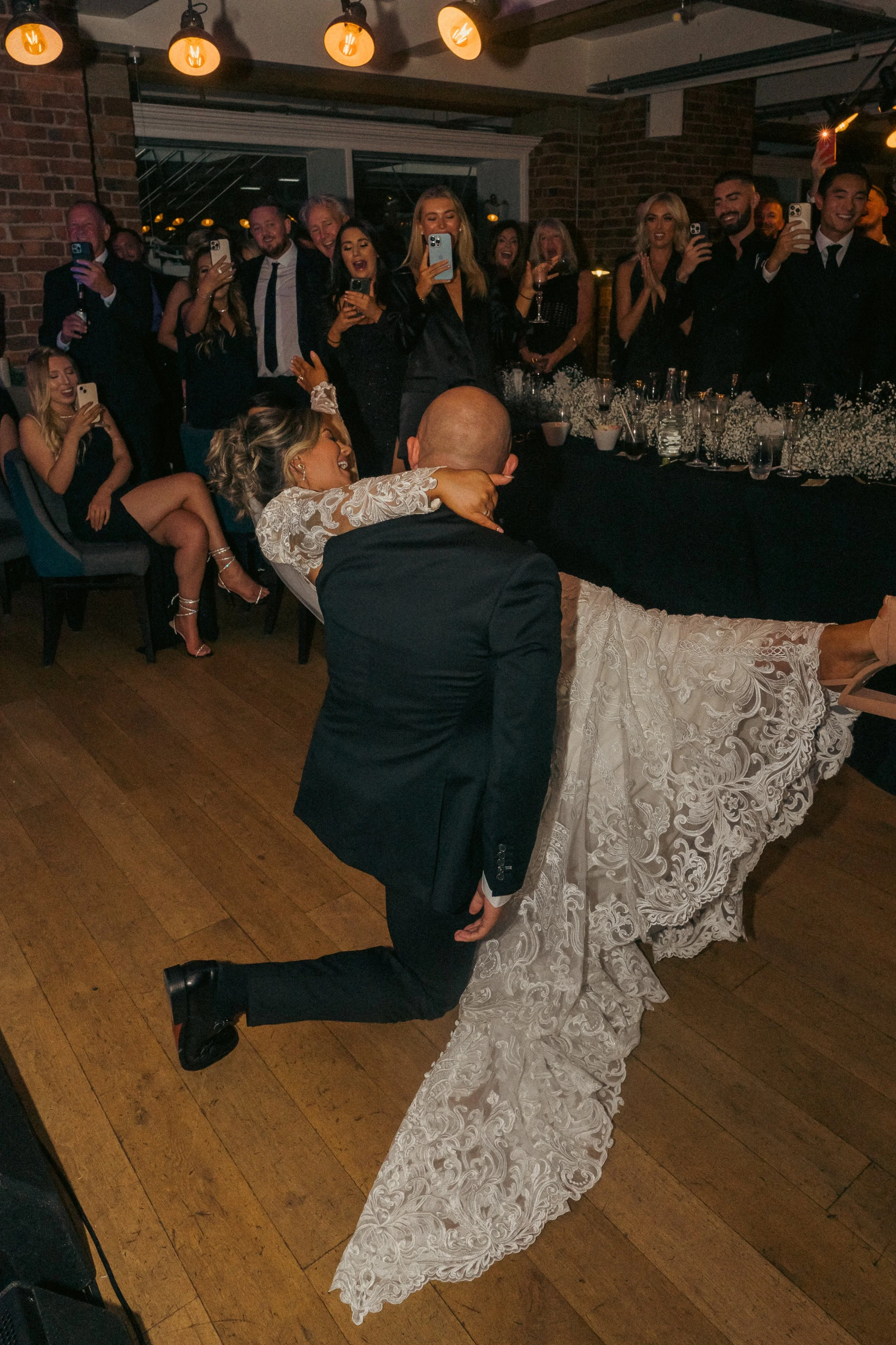 A bride and groom share a kiss during their wedding reception, with guests gathered around them, taking photos and celebrating. The bride wears a lace dress, and the groom is in a black suit.