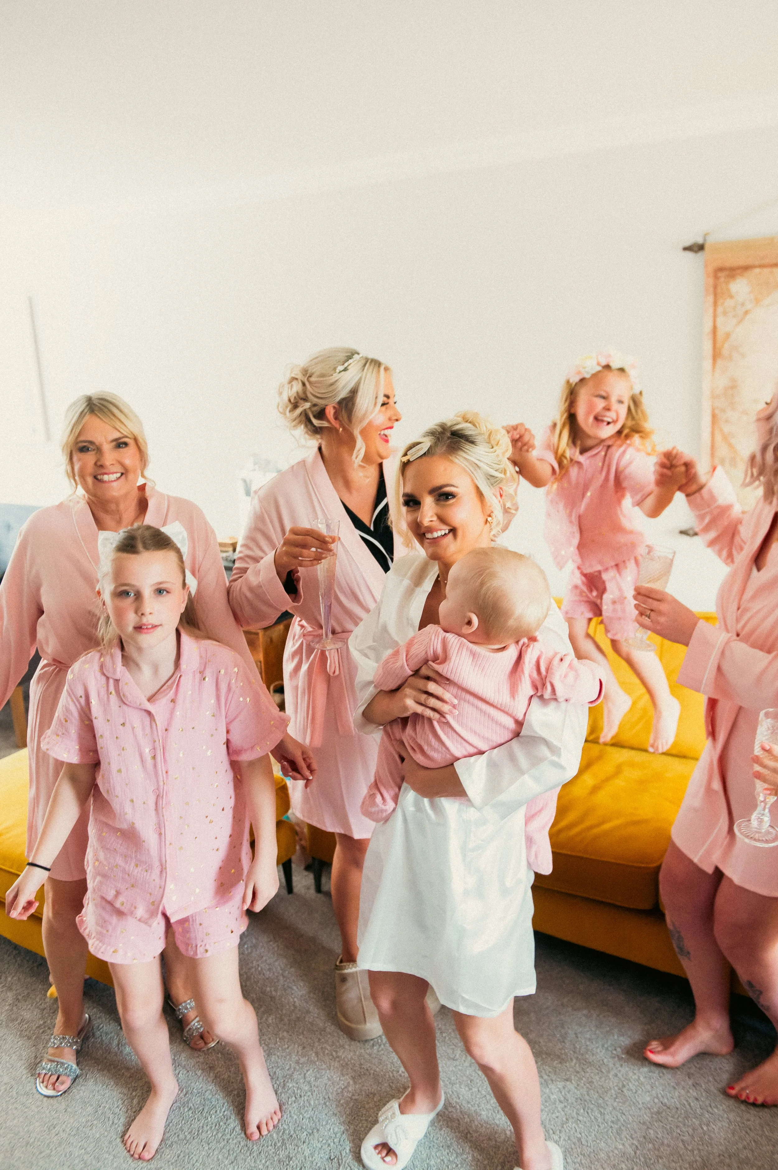 Group of women and children celebrating, dressed in pink and white pajamas, with some holding glasses of champagne in a bright room with a yellow couch.