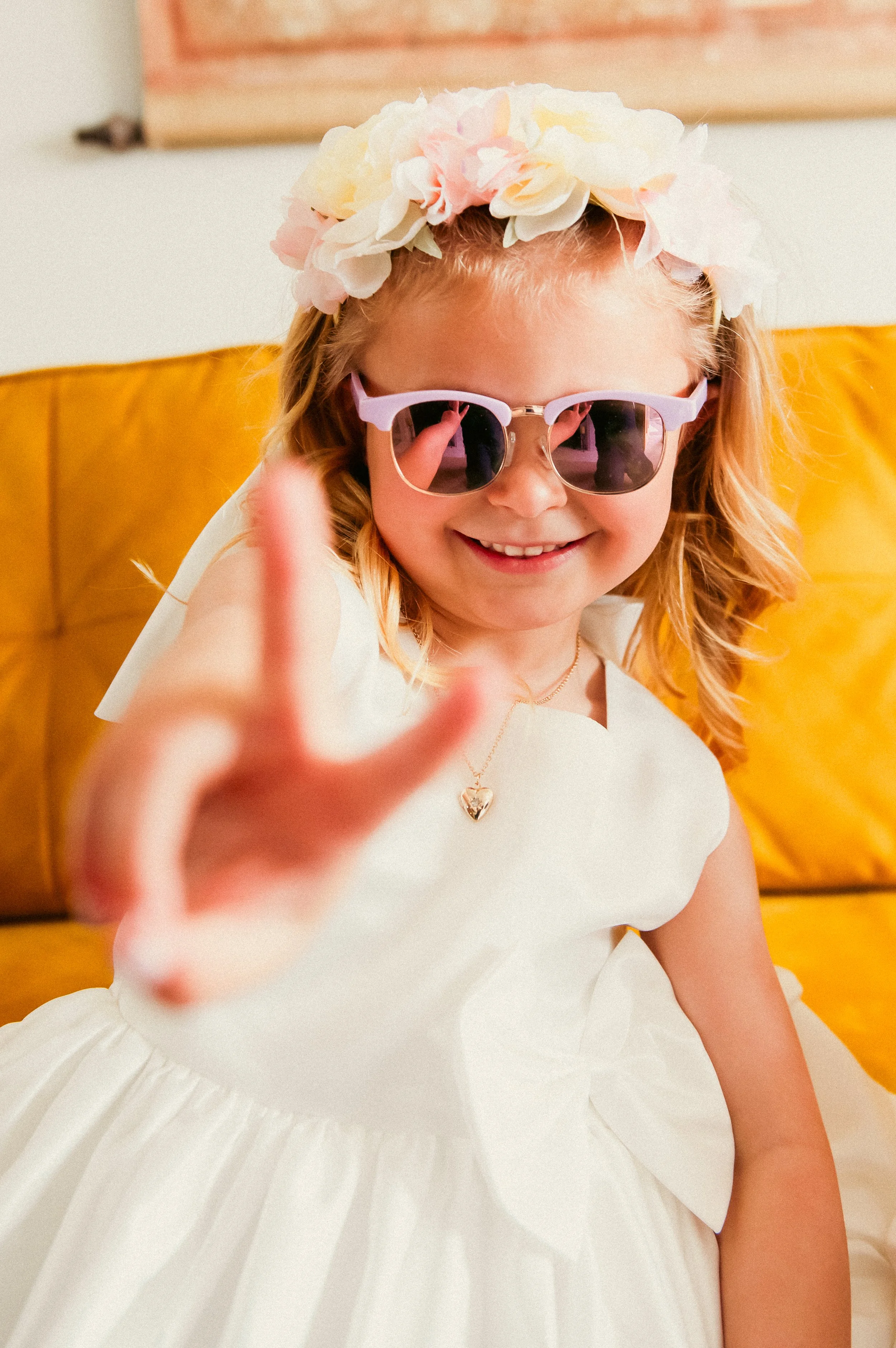 A young girl wearing sunglasses, a flower crown, and a white dress, smiling and reaching towards the camera.
