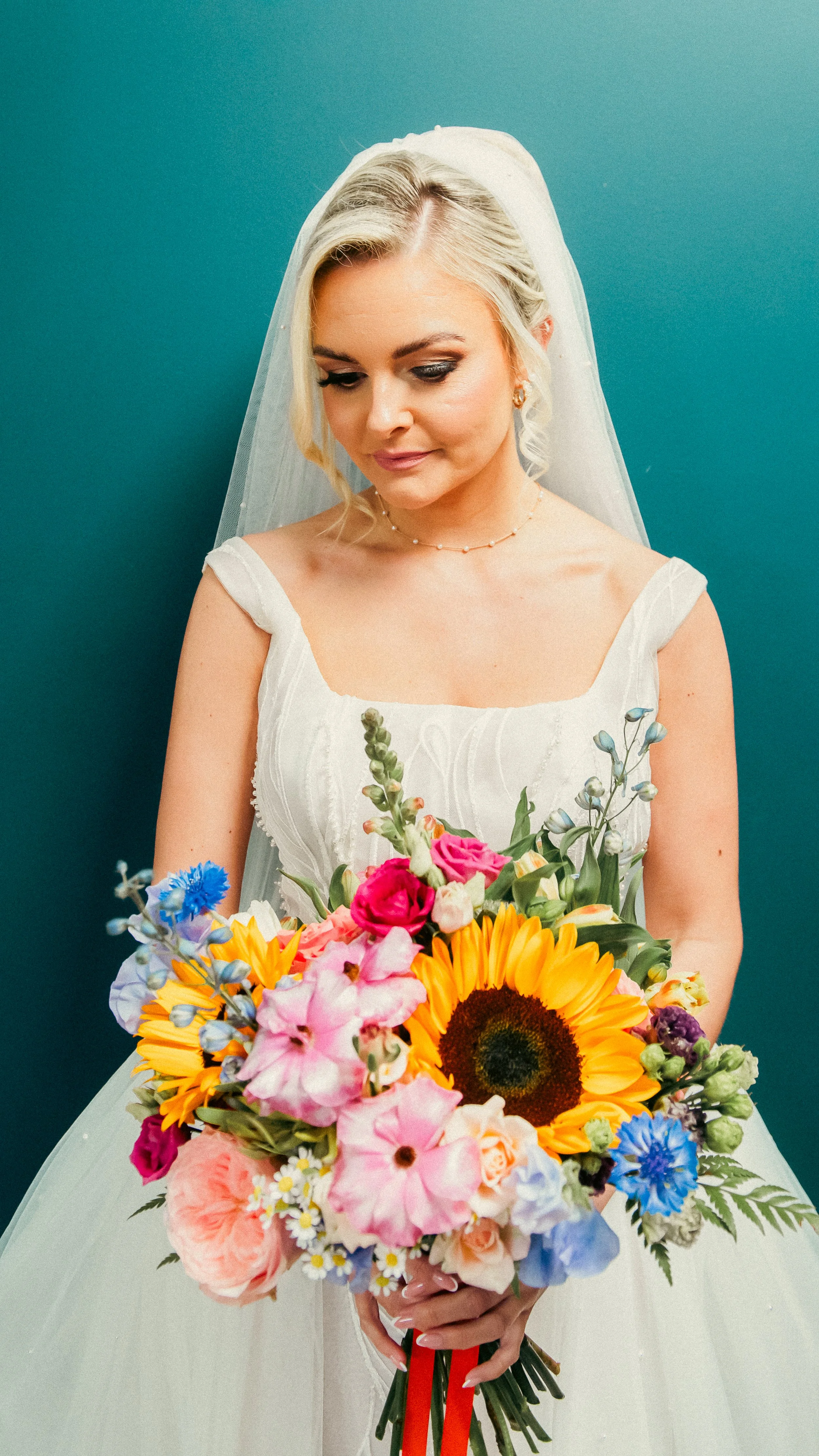 A bride with blonde hair, wearing a white wedding dress and veil, holding a colorful bouquet of flowers, standing against a teal background.