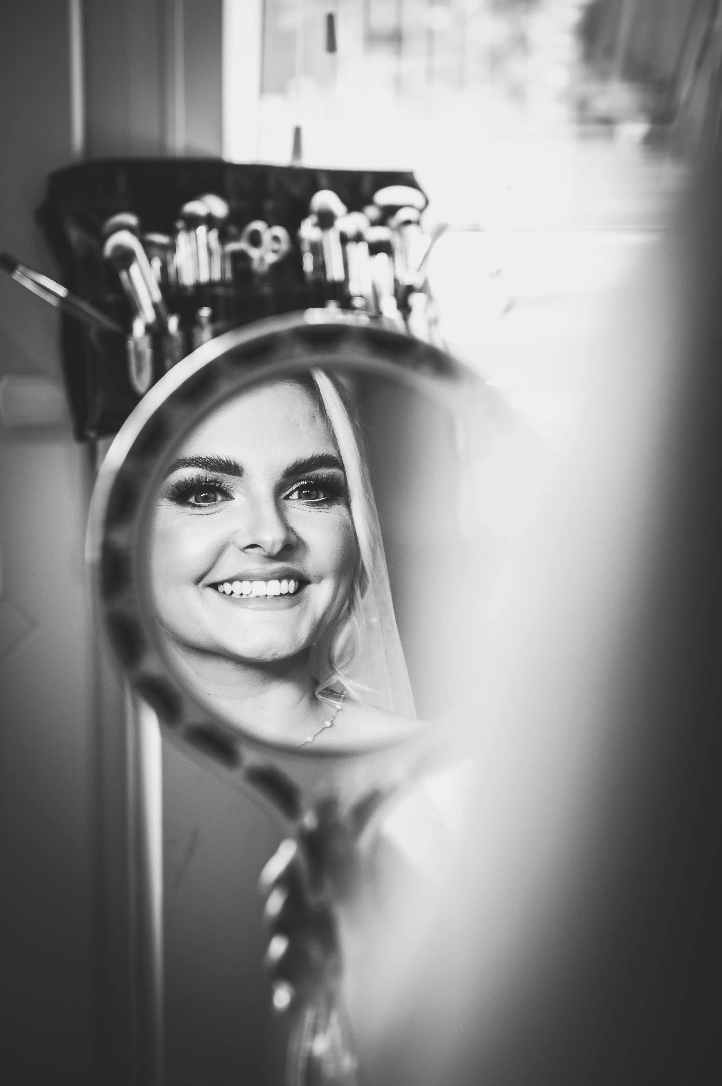 A smiling woman looks into a mirror with makeup brushes behind her, seen through a handheld mirror reflected in a larger mirror.