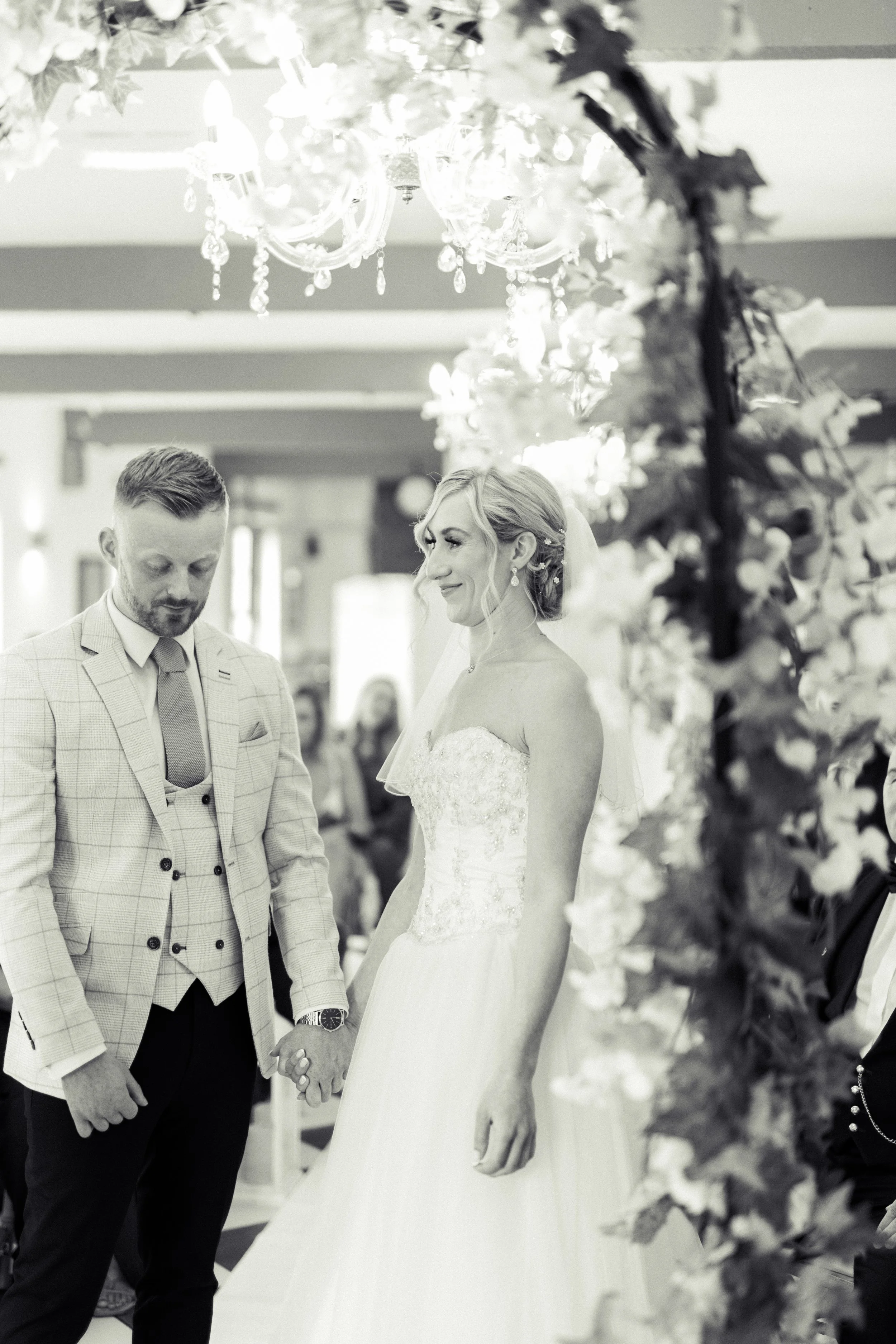 A bride and groom holding hands during their wedding ceremony indoors, with floral decorations and a chandelier overhead.