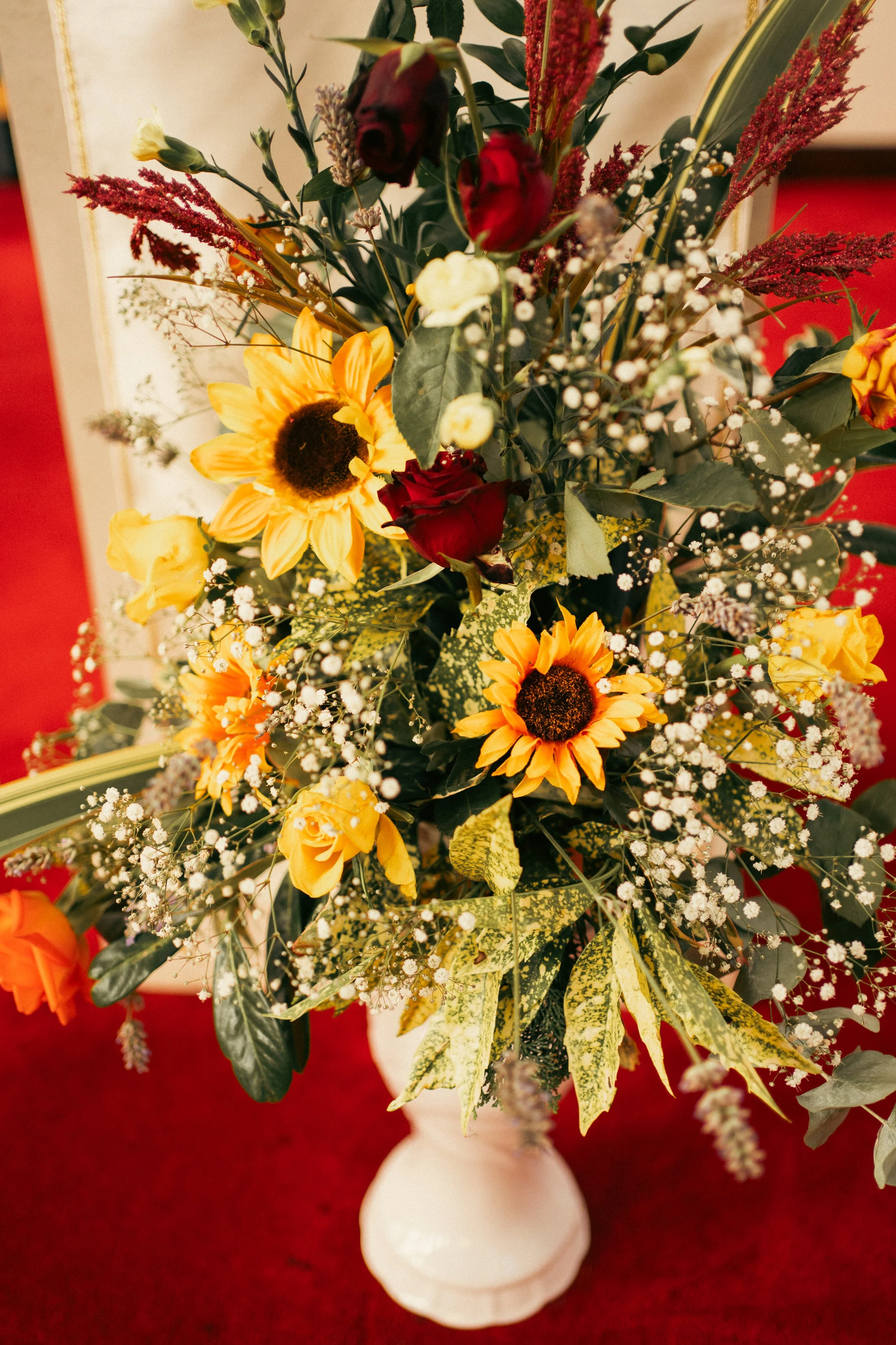 A bouquet of yellow sunflowers, red roses, and white baby's breath in a white vase on a red surface.