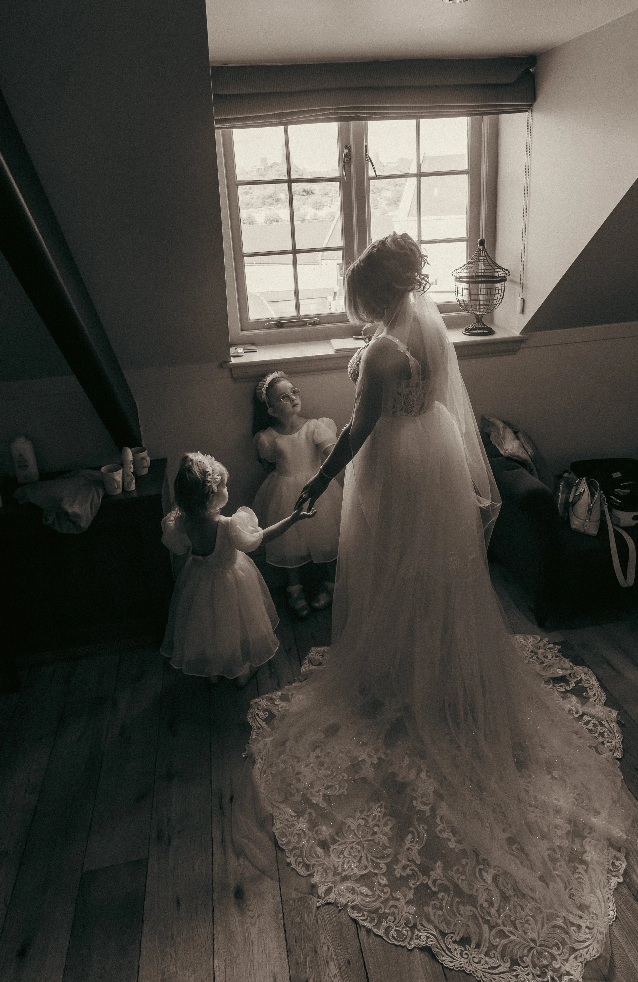 A bride in a wedding dress holding hands with two young girls in a room with wooden flooring and a window. The girls are wearing white dresses and headbands, and the bride is wearing a lace gown with a long train and veil.