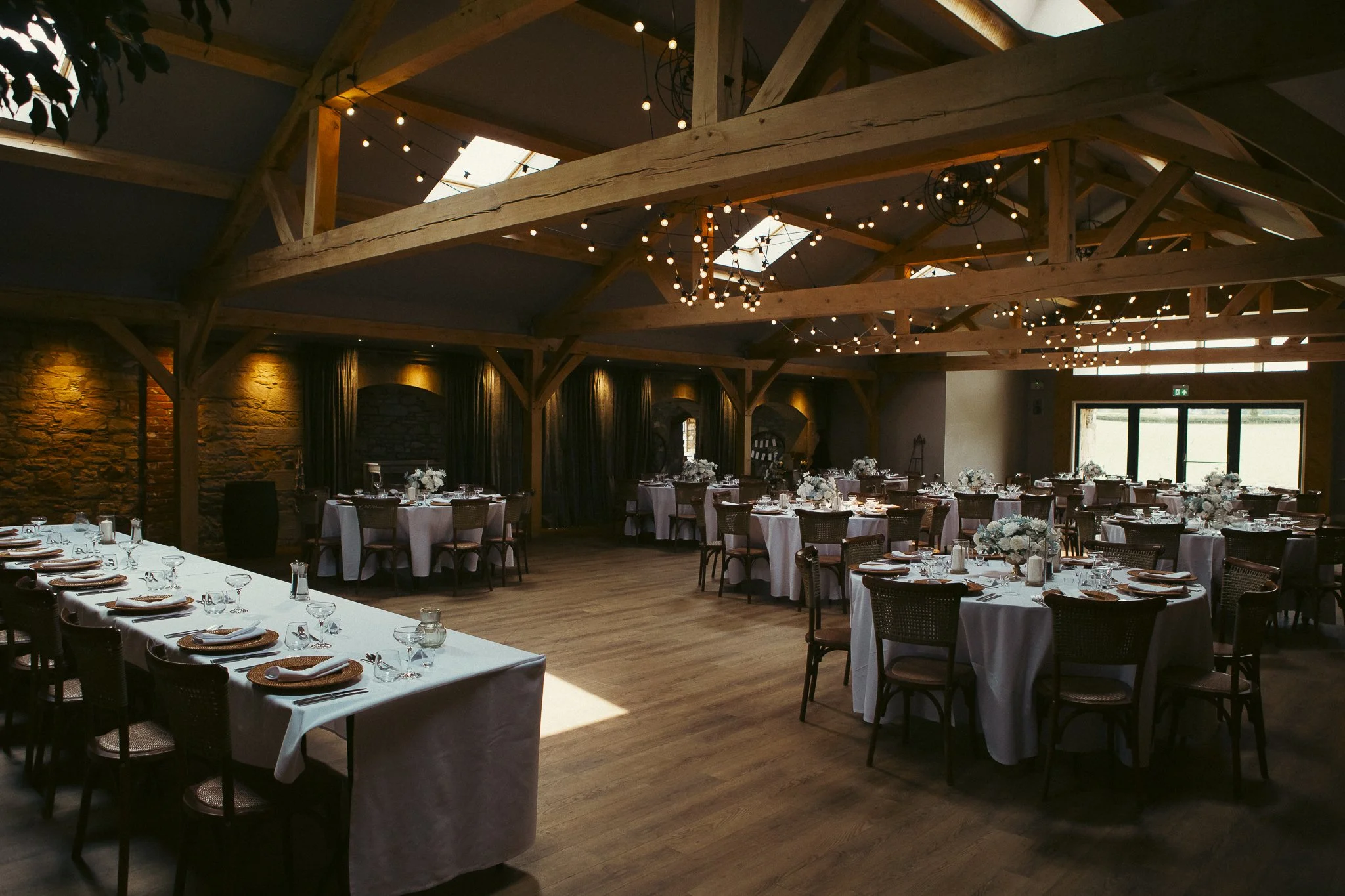 A spacious event hall with wooden beams and string lights, decorated with round tables covered in white tablecloths, floral centerpieces, and place settings, set up for a formal gathering or celebration.