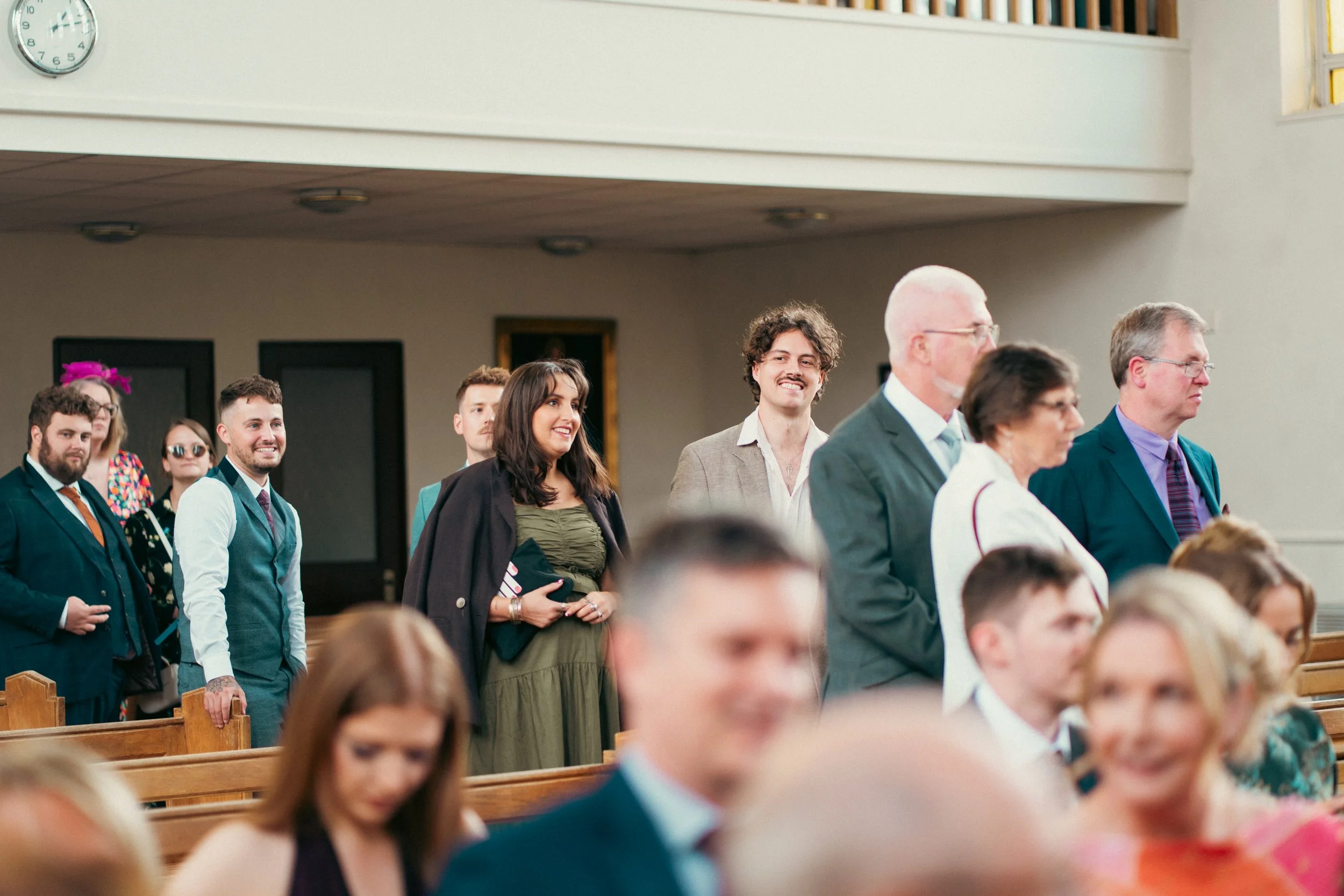People standing and sitting in pews inside a church, attending a formal event, with some smiling and others looking attentive.